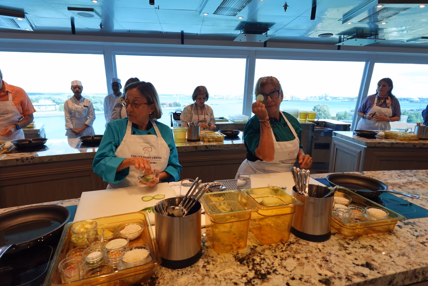 Two women in aprons preparing cucumber slices in a cooking class kitchen with other participants in the background and a waterfront view.