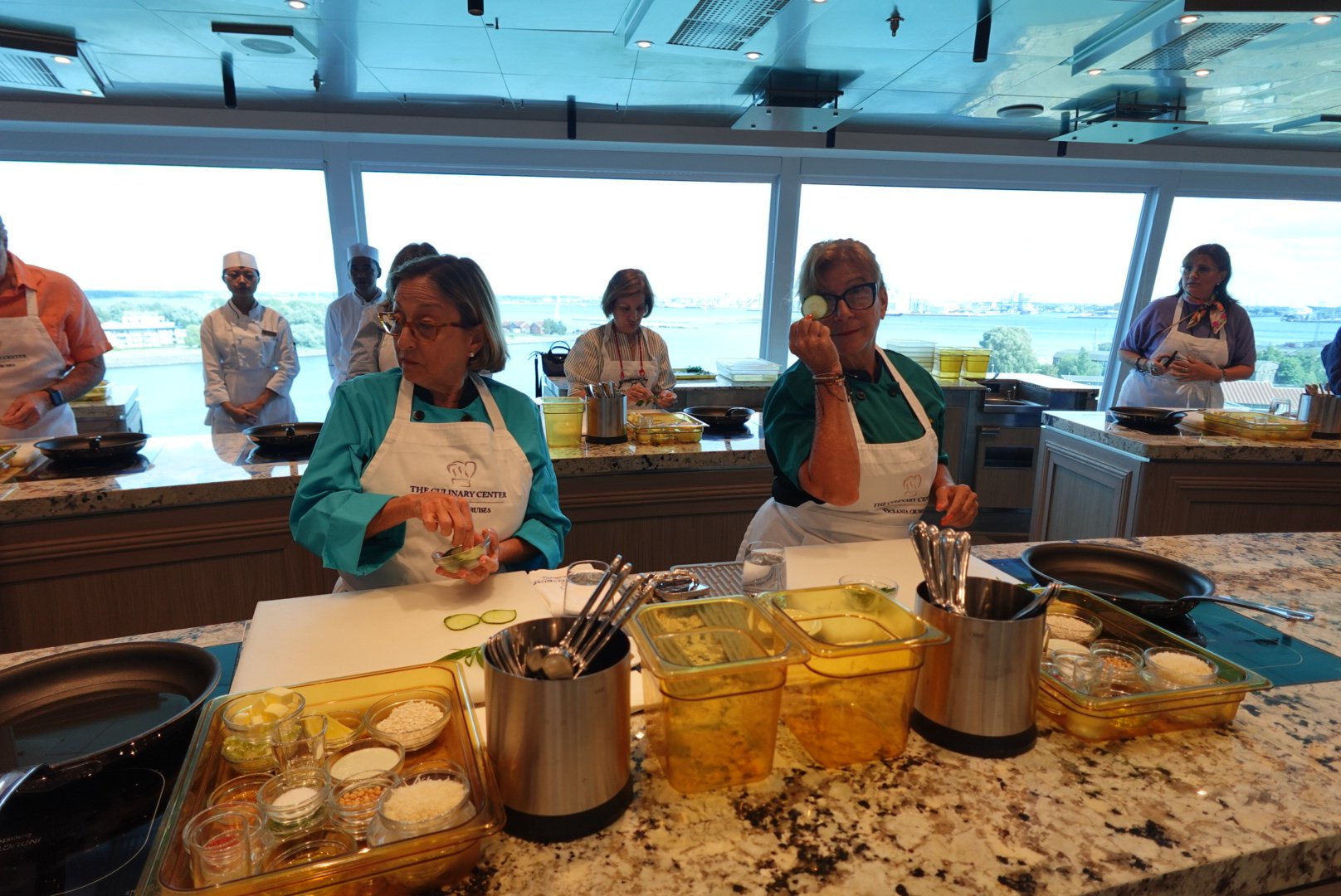 Two women in aprons preparing cucumber slices in a cooking class kitchen with other participants in the background and a waterfront view.