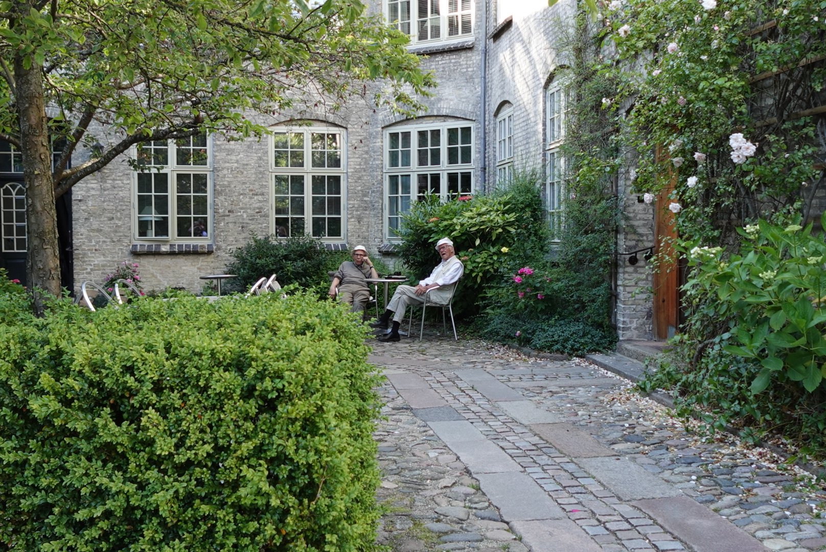 Two elderly men sitting on chairs chatting in a green garden courtyard with brick walls and windows in the background.