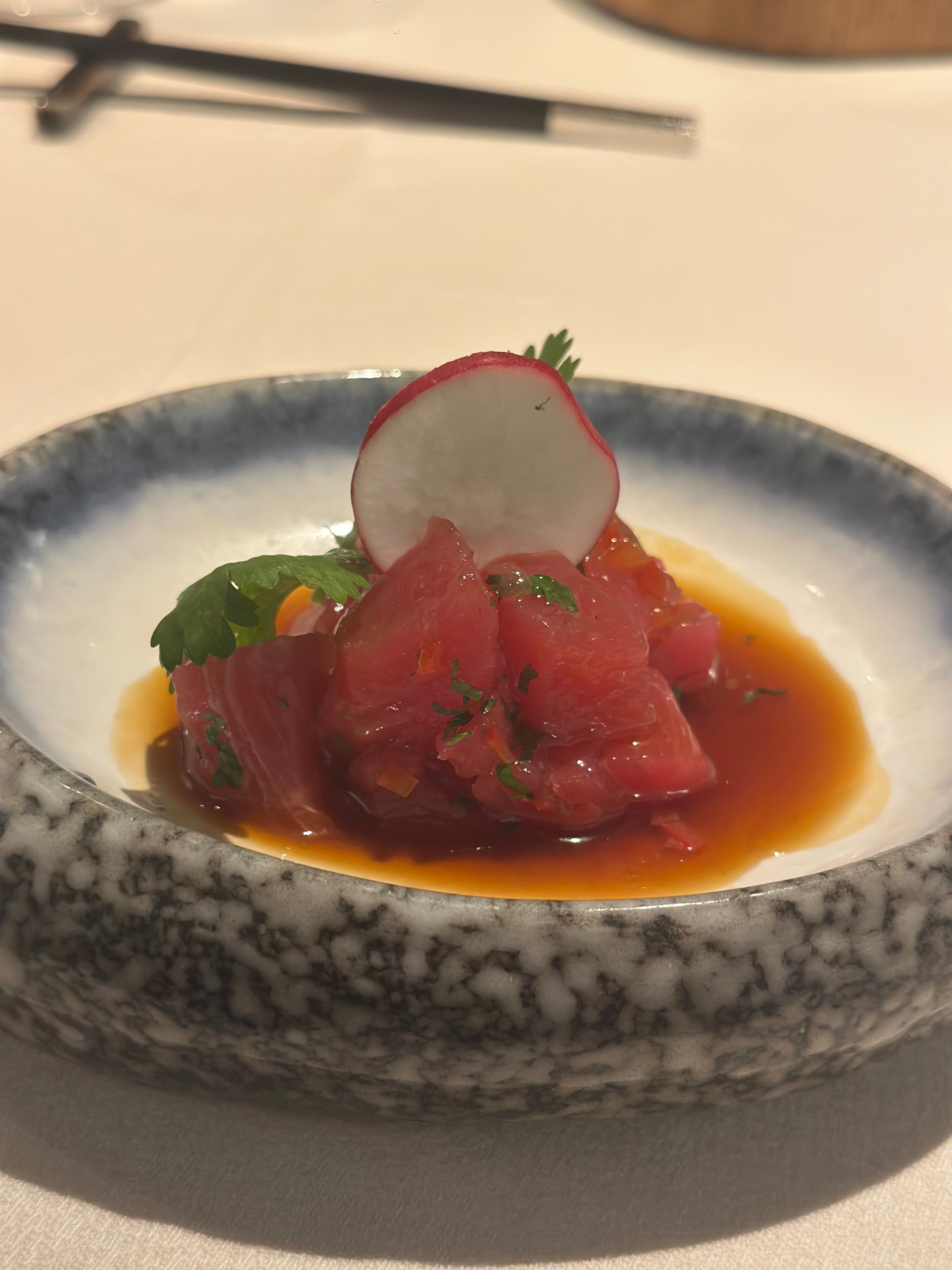 Diced tuna sashimi served in a grey marbled bowl with orange sauce, garnished with a radish slice and green herbs.