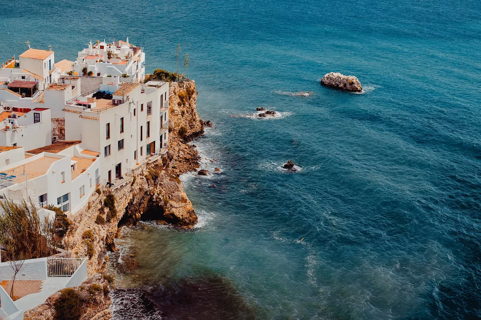 White buildings perched on a rocky seaside cliff overlooking clear blue ocean water with scattered rocks.