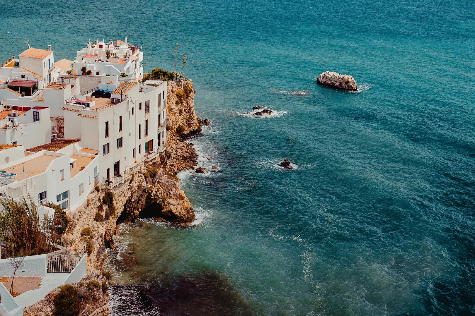 White buildings perched on a rocky seaside cliff overlooking clear blue ocean water with scattered rocks.