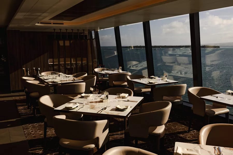 Interior of a restaurant with beige chairs and tables set with glasses and cutlery, overlooking a large body of water through floor-to-ceiling windows.