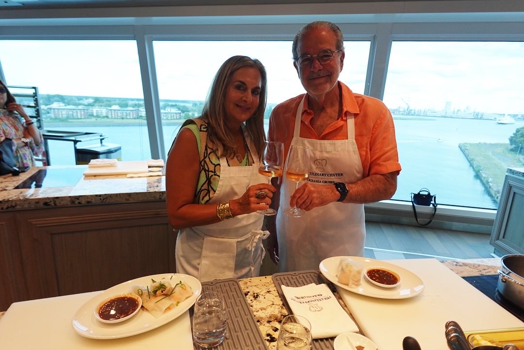 A man and woman wearing aprons holding wine glasses and smiling in a kitchen with a waterfront view.