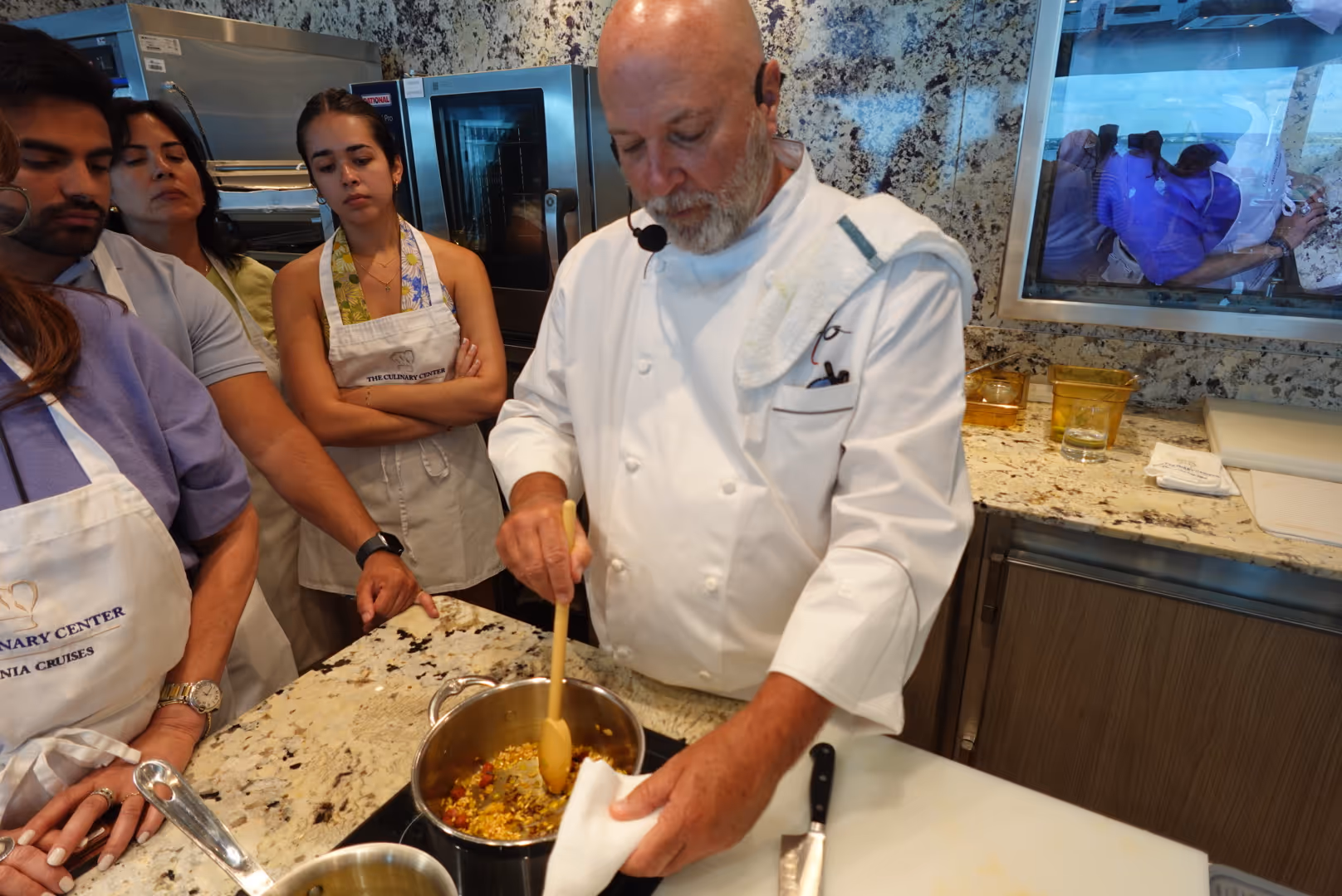 Chef in white coat stirring food in a pot while a group of people watch attentively in a kitchen setting.