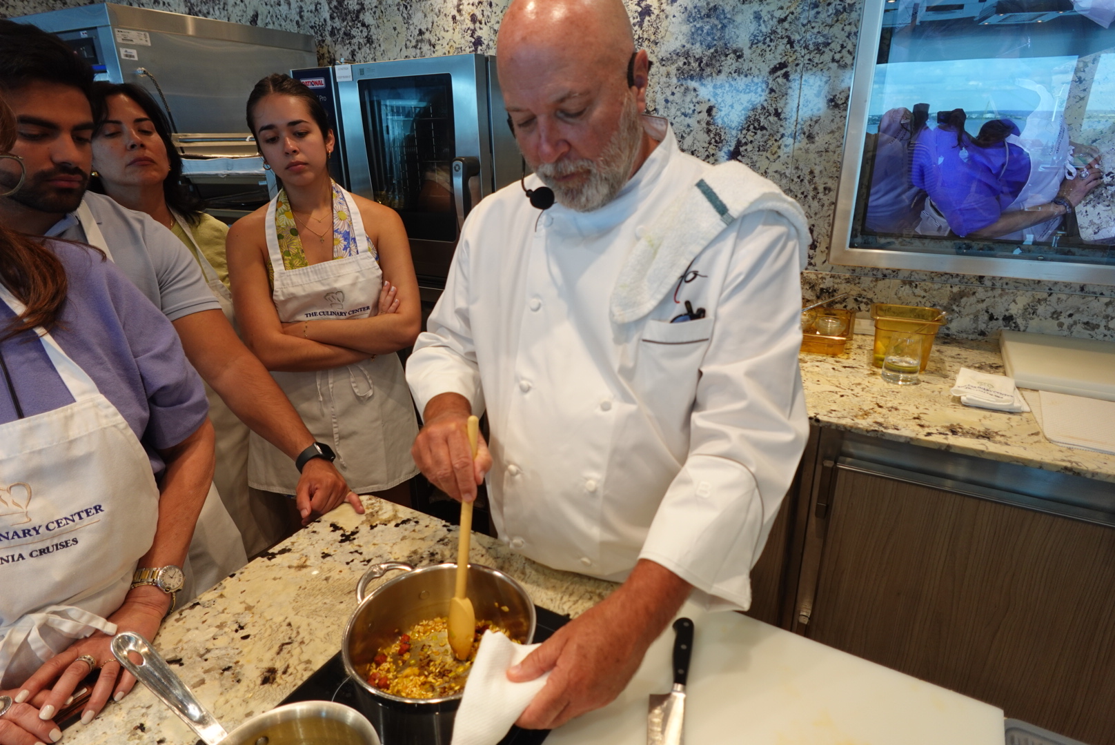 Chef in white coat stirring food in a pot while a group of people watch attentively in a kitchen setting.