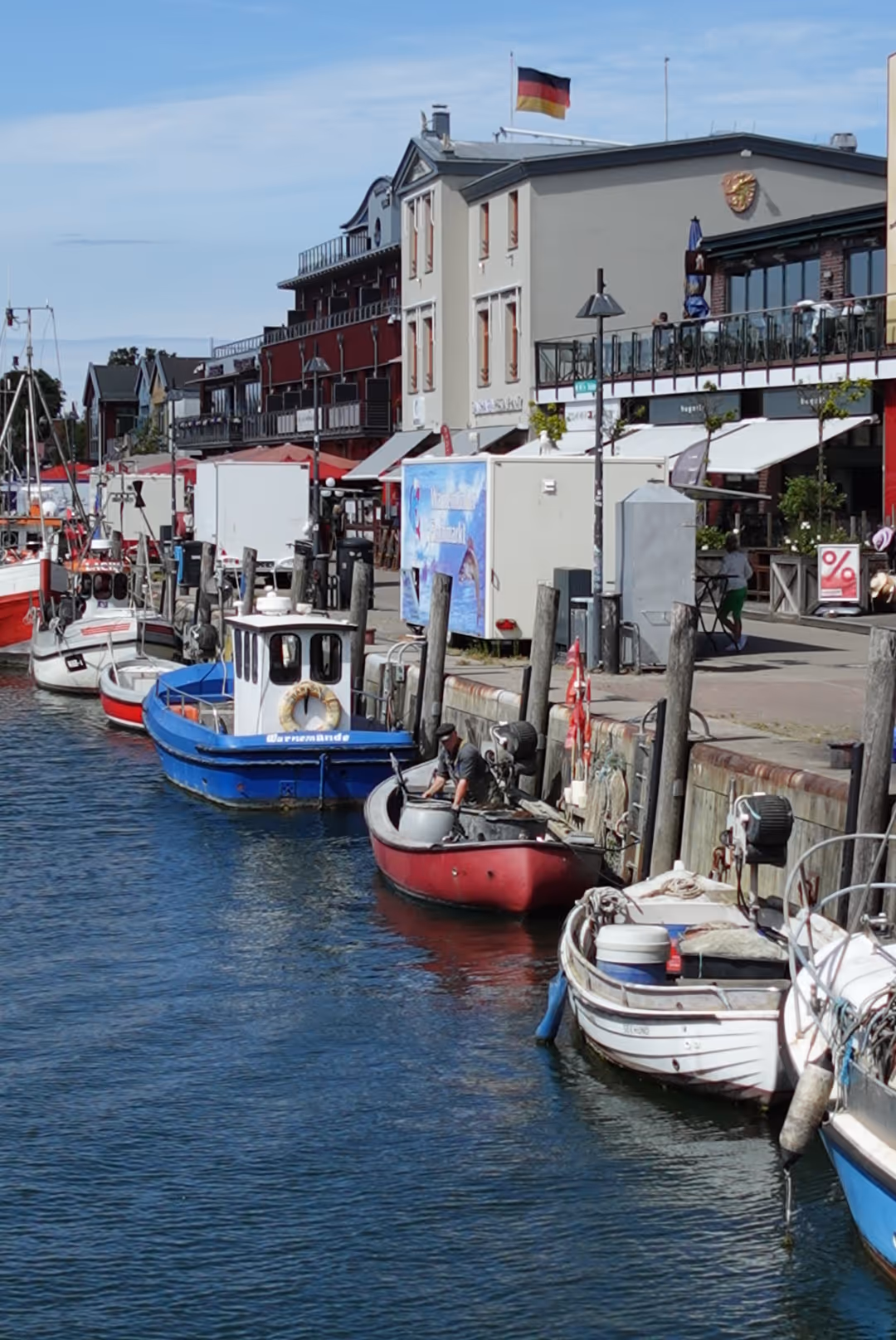 Small boats docked along a waterfront with buildings and a German flag in the background under a partly cloudy sky.