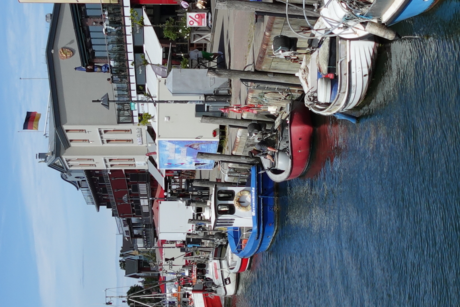 Small boats docked along a waterfront with buildings and a German flag in the background under a partly cloudy sky.