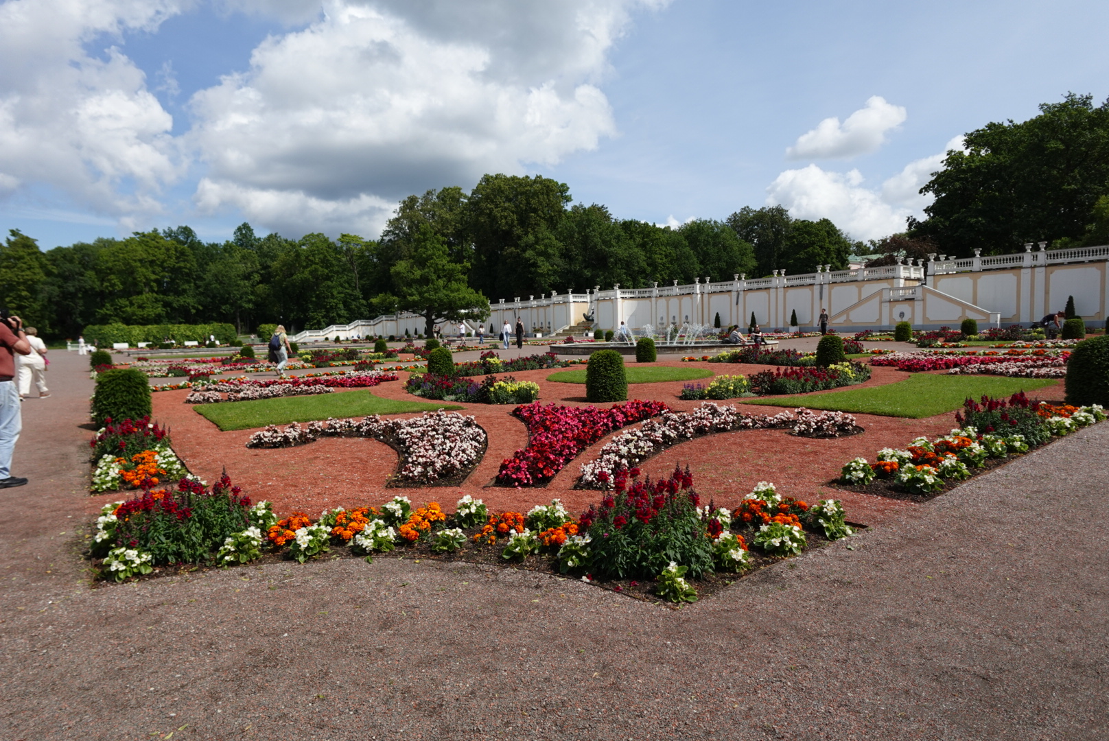 Formal garden with colorful flower beds, green shrubs, and a white balustrade wall under a partly cloudy sky.