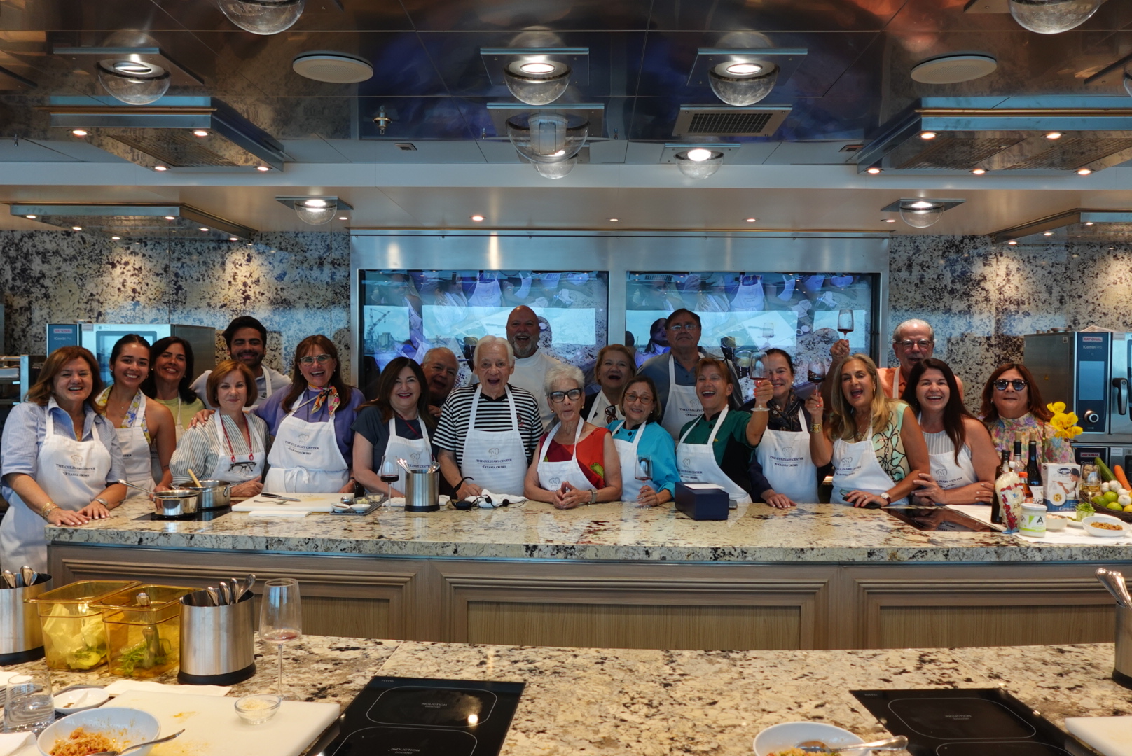 Group of adults wearing aprons standing behind a kitchen counter in a cooking class setting, smiling and holding wine glasses.
