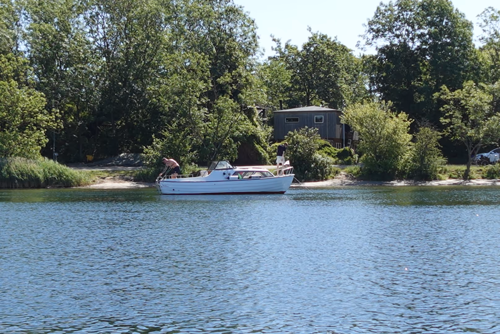 Two people on a small white motorboat near a tree-lined shore with a cabin and a parked car in the background.