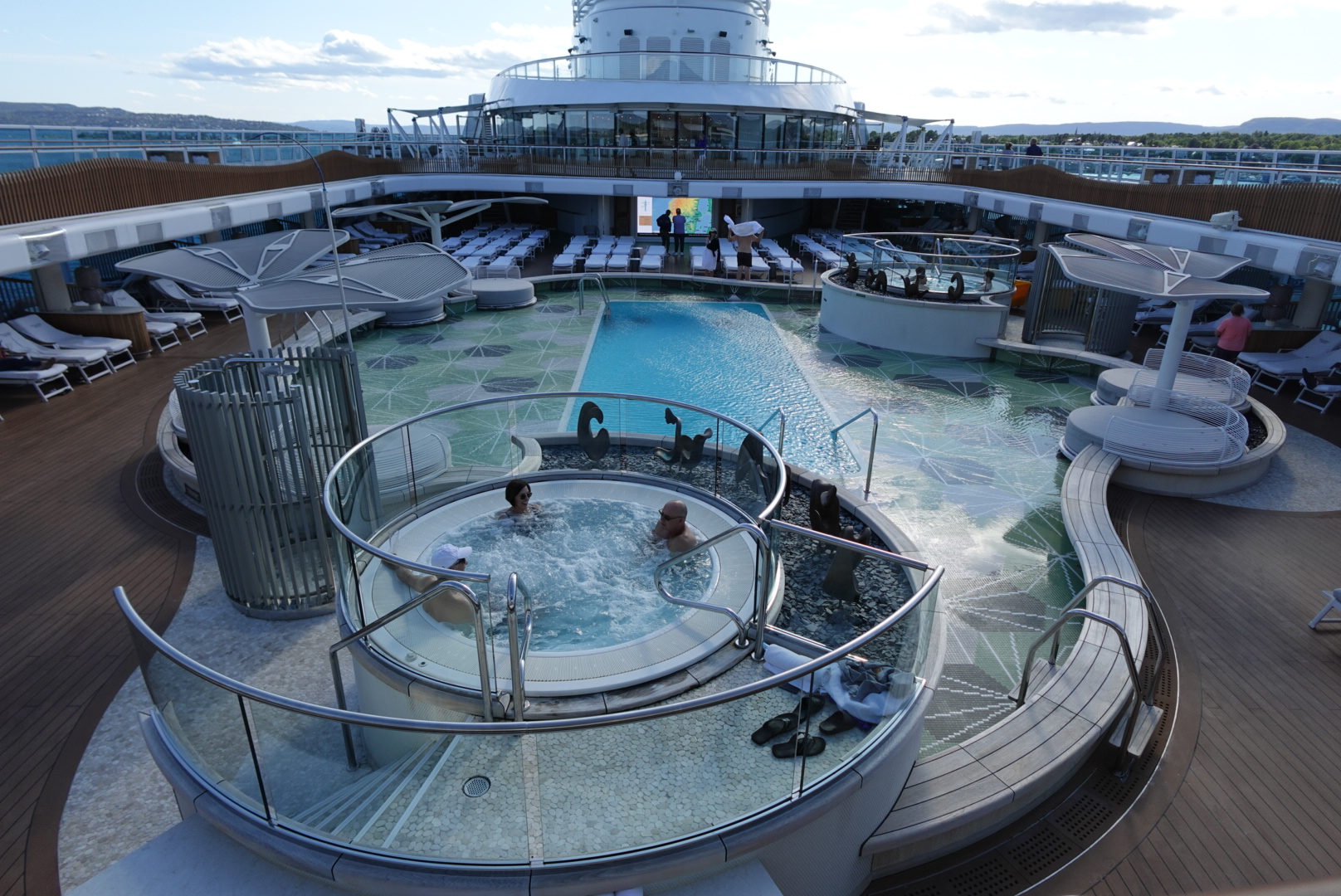 Three people relaxing in a circular hot tub on a cruise ship deck with a pool, lounge chairs, and cityscape in the background.