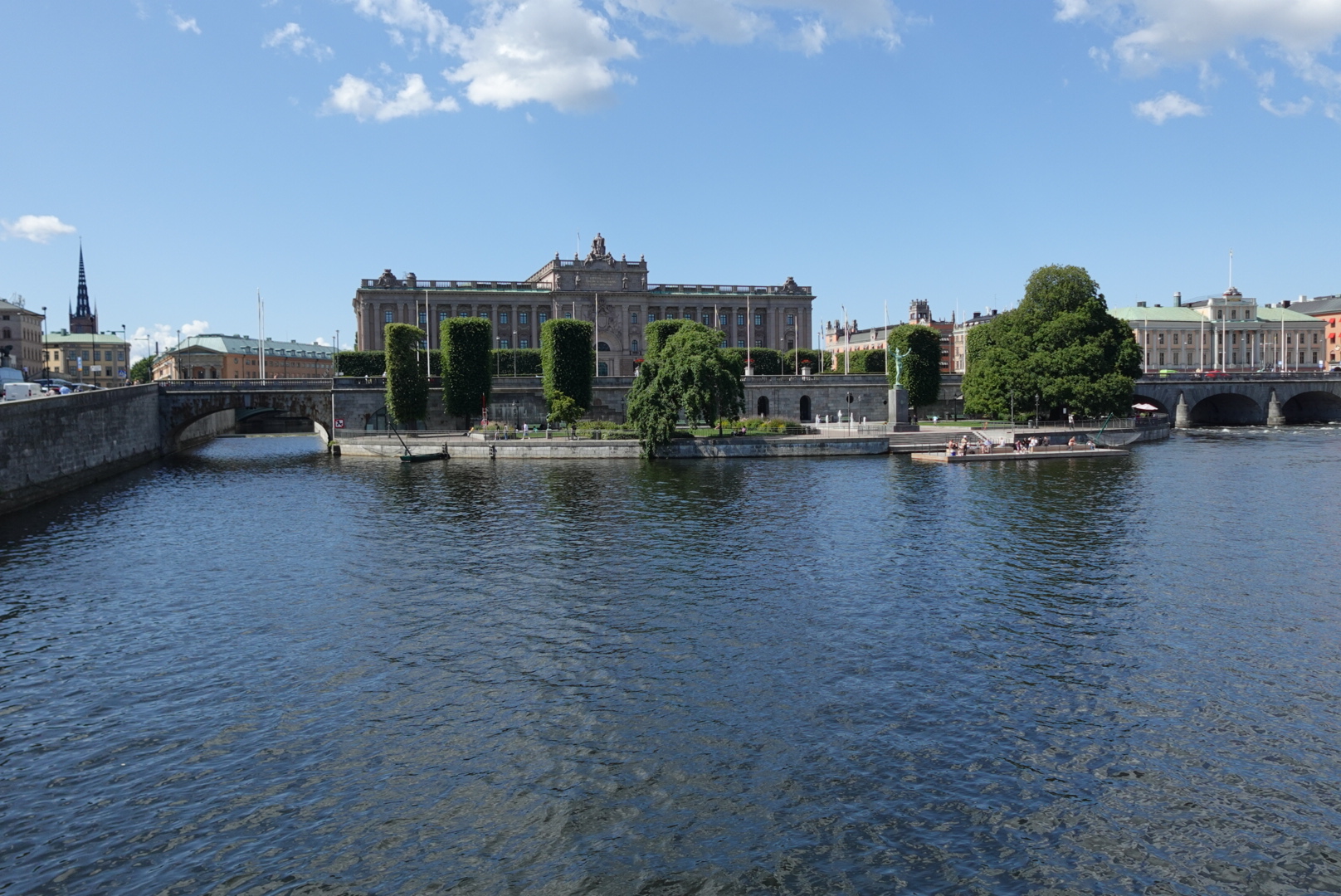 Large classical building with columns behind river and green trees under a blue sky with scattered clouds.