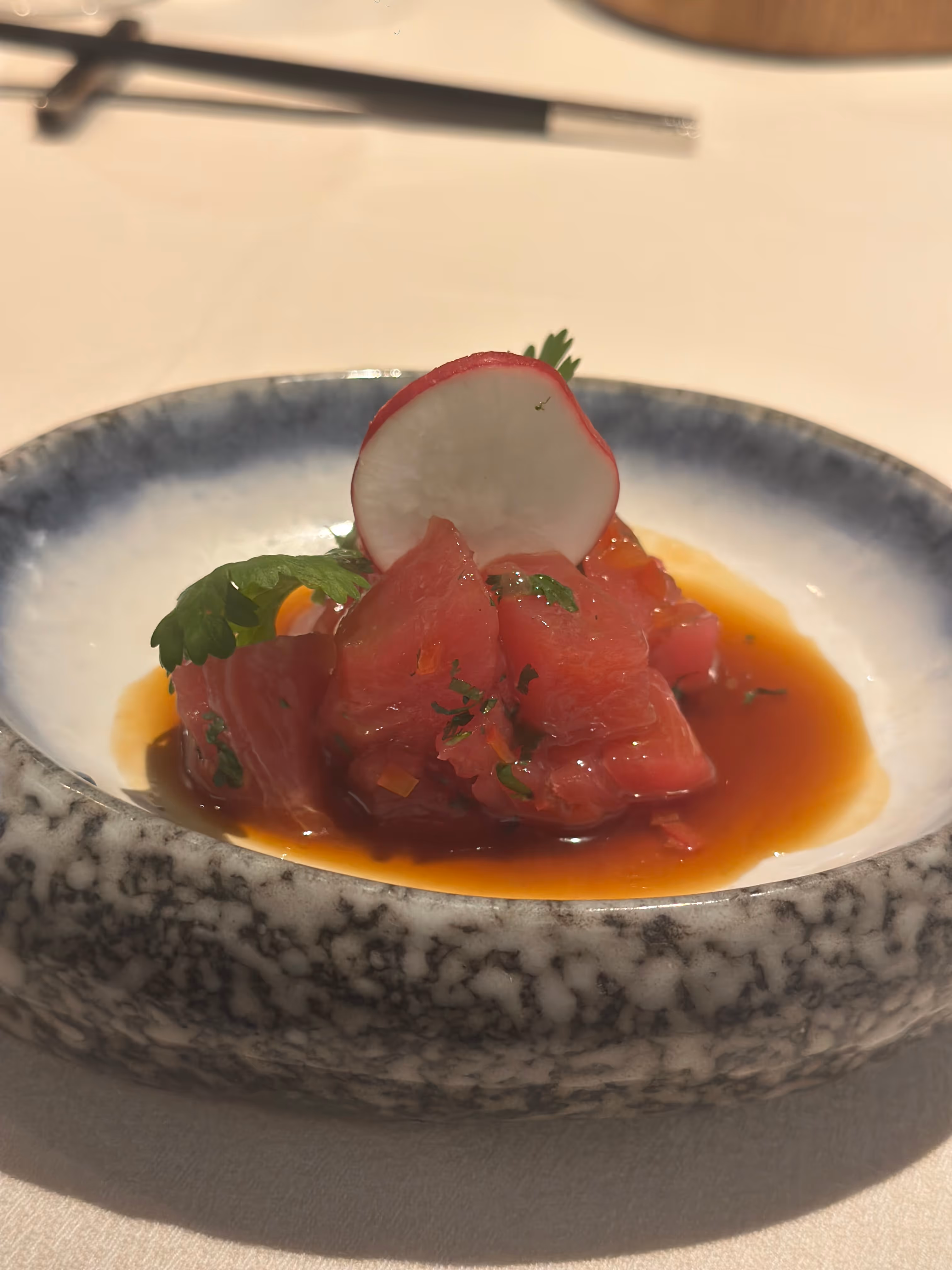 A dish of diced raw tuna topped with a radish slice and herbs, served in a shallow bowl with a brown sauce.