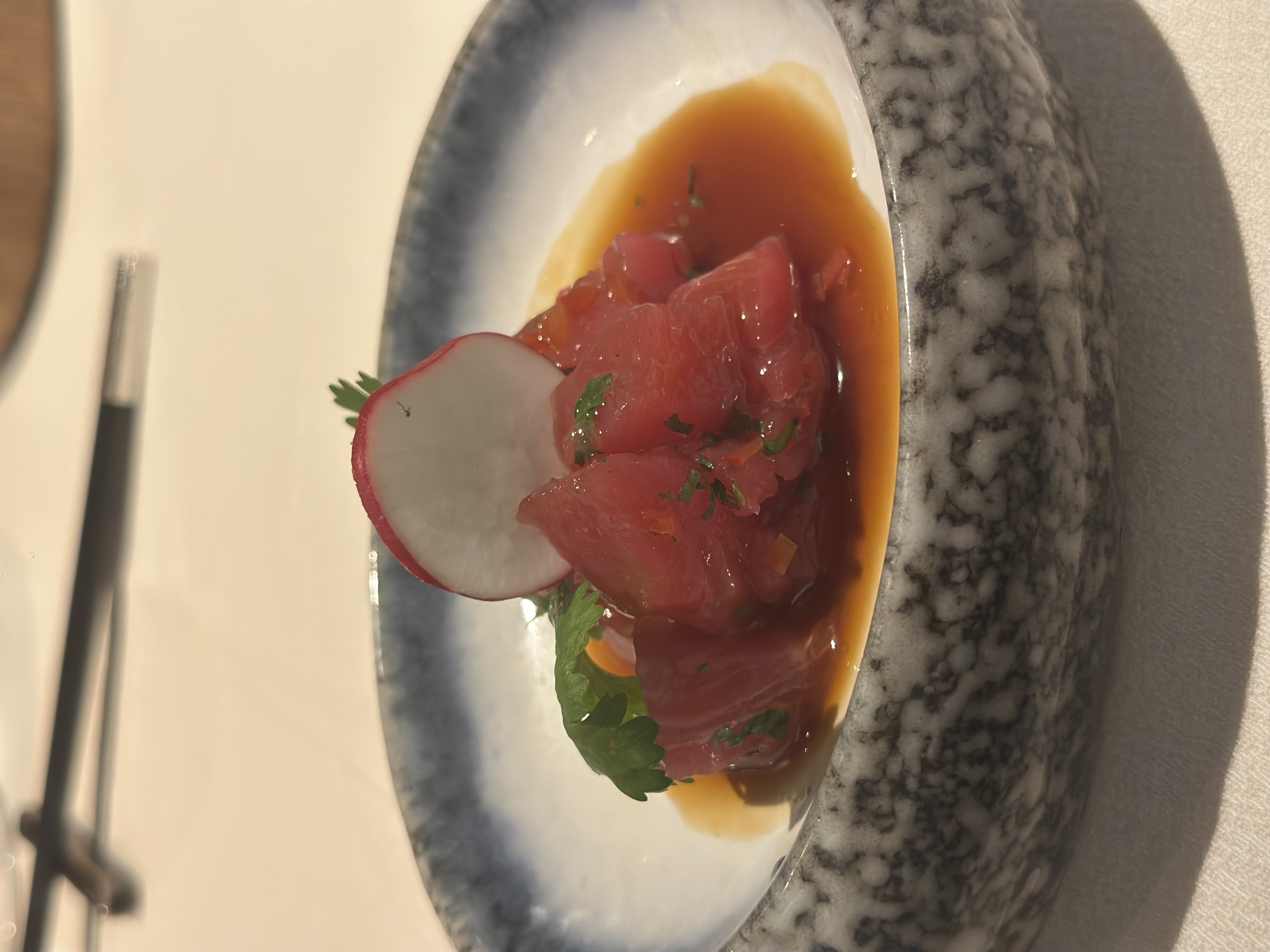 A dish of diced raw tuna topped with a radish slice and herbs, served in a shallow bowl with a brown sauce.