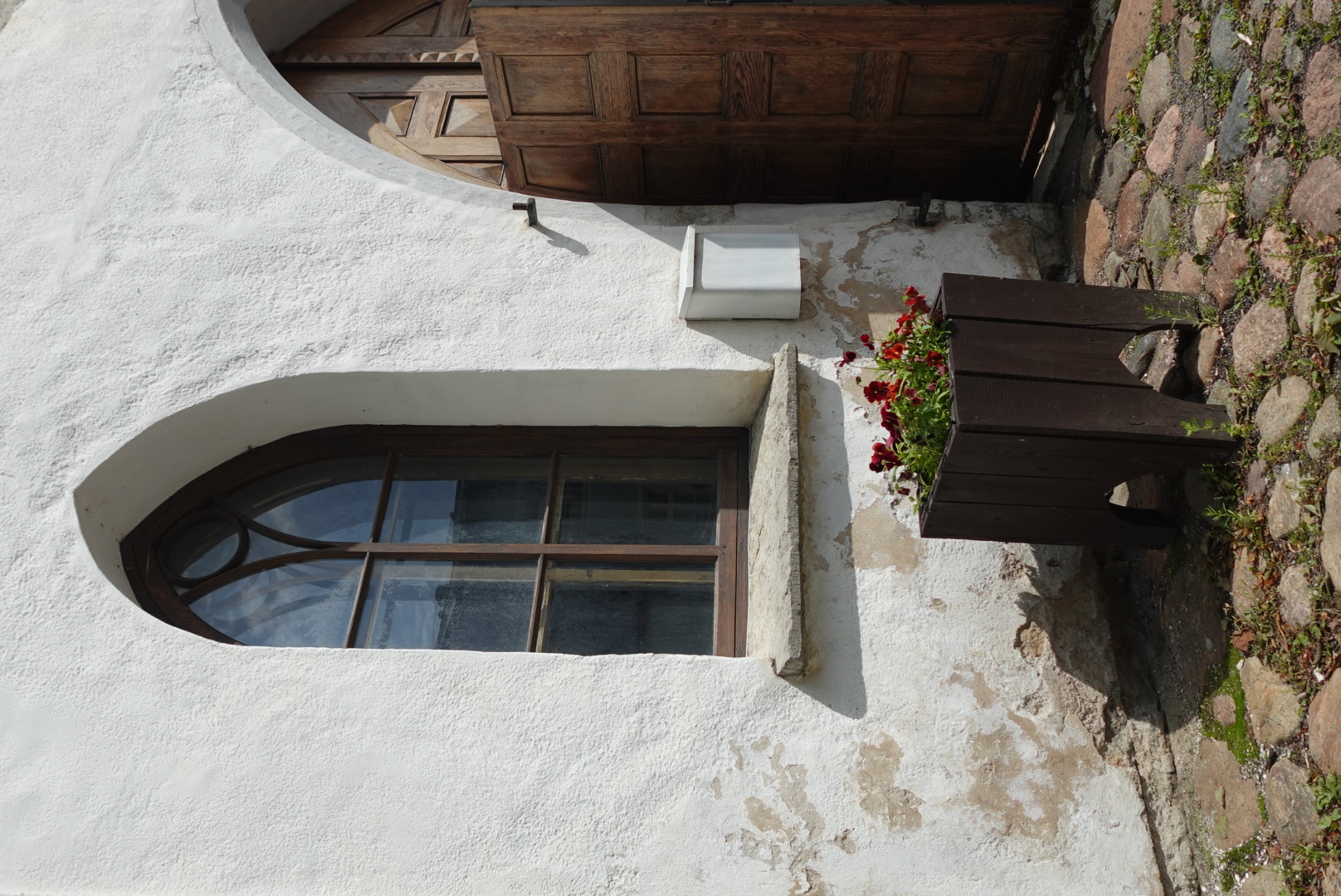 Arched wooden window and partially open wooden door on a white textured wall with a dark wooden planter box filled with red flowers on a cobblestone ground.
