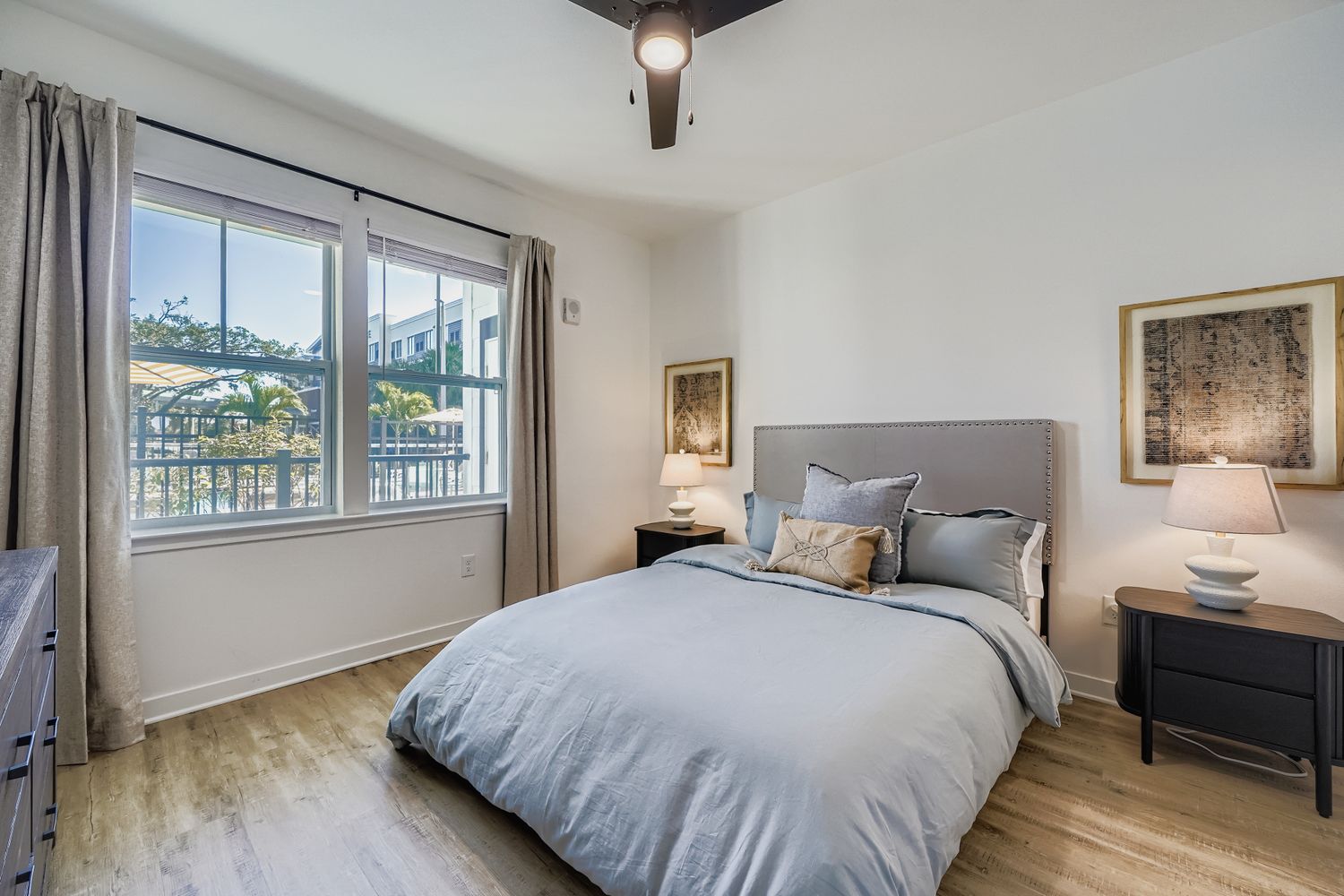 An apartment bedroom with light wood-style flooring, a ceiling fan, a queen-sized bed with a tufted headboard, and a large window overlooking an outdoor area.