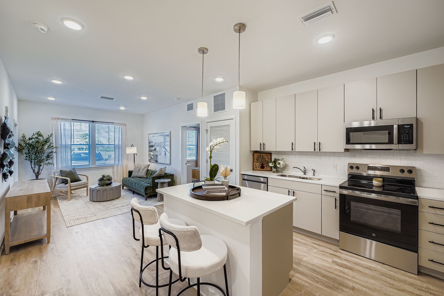 An open-concept living area and kitchen featuring a white center island with pendant lighting, light cabinets, stainless steel appliances, and a living space with a large window.