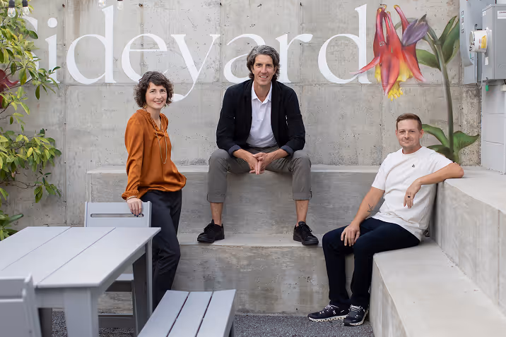 Three people posing casually on concrete steps and benches with a large flower mural and partial text 'ideyard' in the background.
