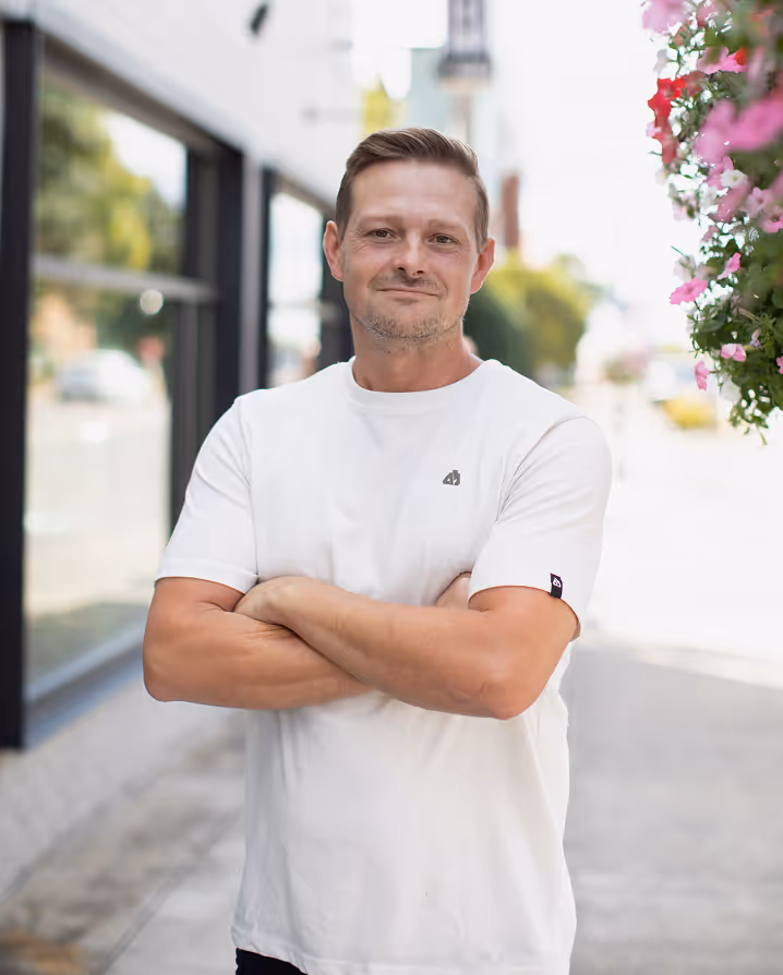 Smiling man in a white t-shirt standing outdoors with arms crossed, with flowers and buildings in the background.