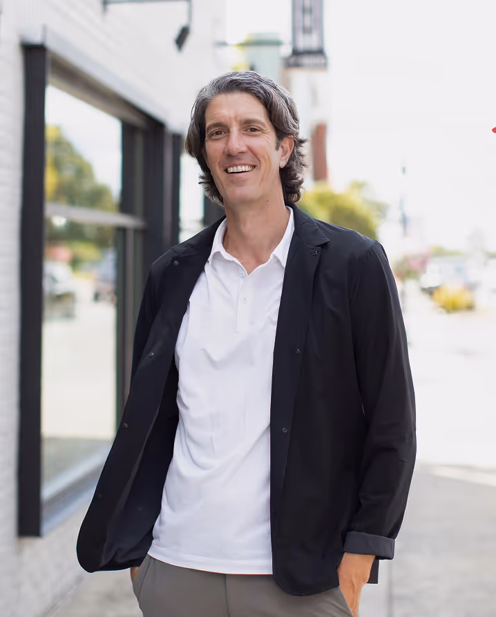 Smiling man with gray hair wearing a white shirt and black jacket standing outdoors on a city sidewalk.