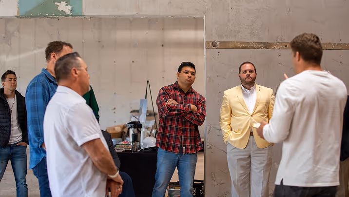 Group of men standing indoors engaged in a serious discussion, with one man in a yellow blazer and another speaking in front.