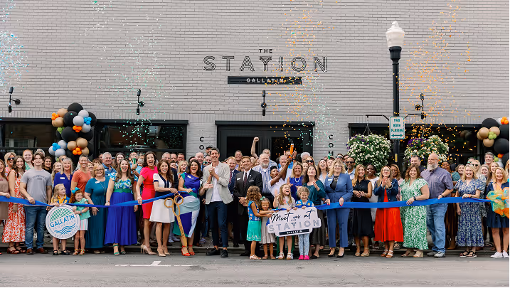 A large group of people gathered in front of The Station Gallatin for a ribbon-cutting ceremony with balloons and confetti.