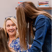 Smiling woman with glasses on her head looking up at a younger woman with long hair in a denim jacket.