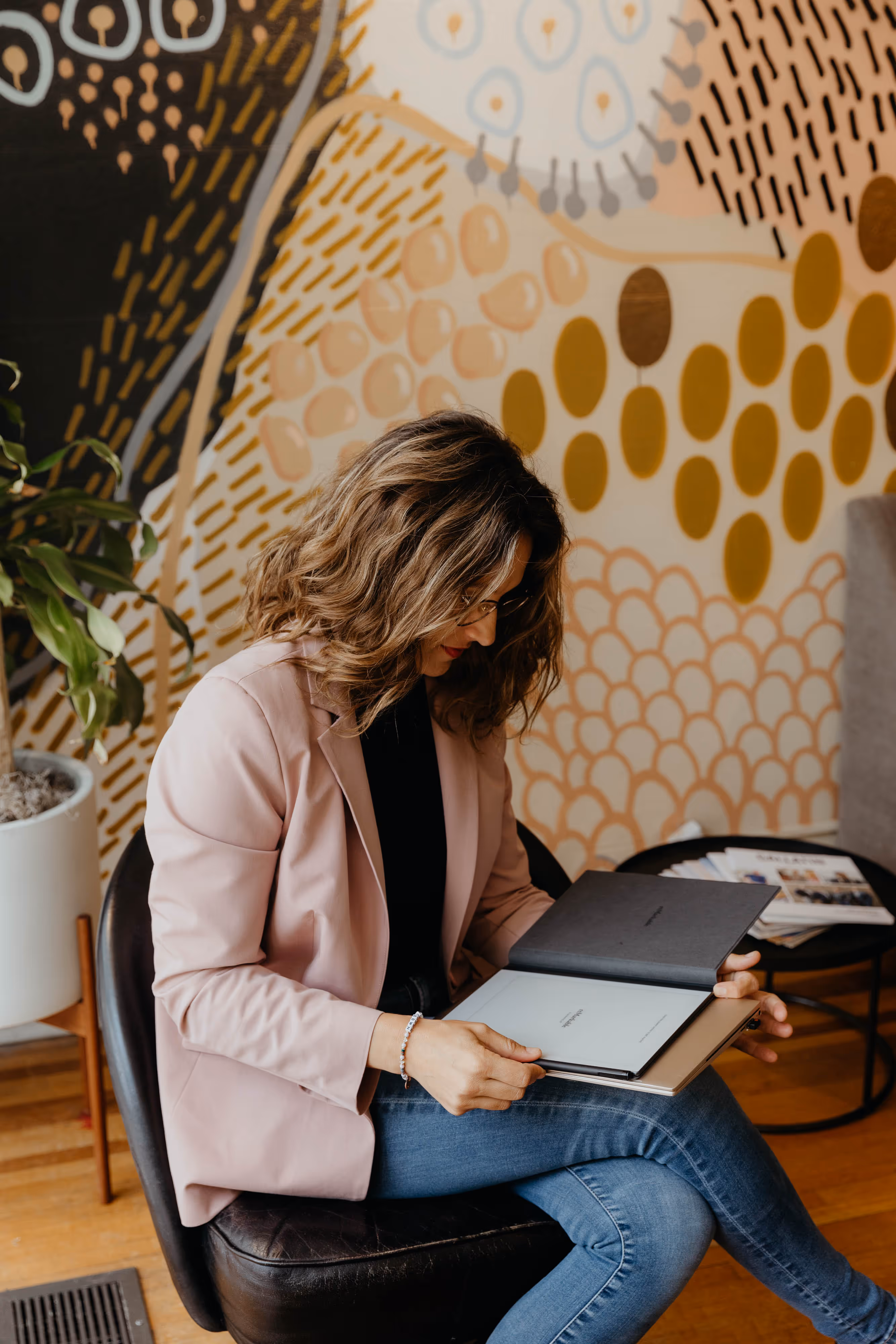 Woman with wavy hair wearing a pink blazer and glasses sitting on a chair reading a book in a room with colorful abstract wall art.