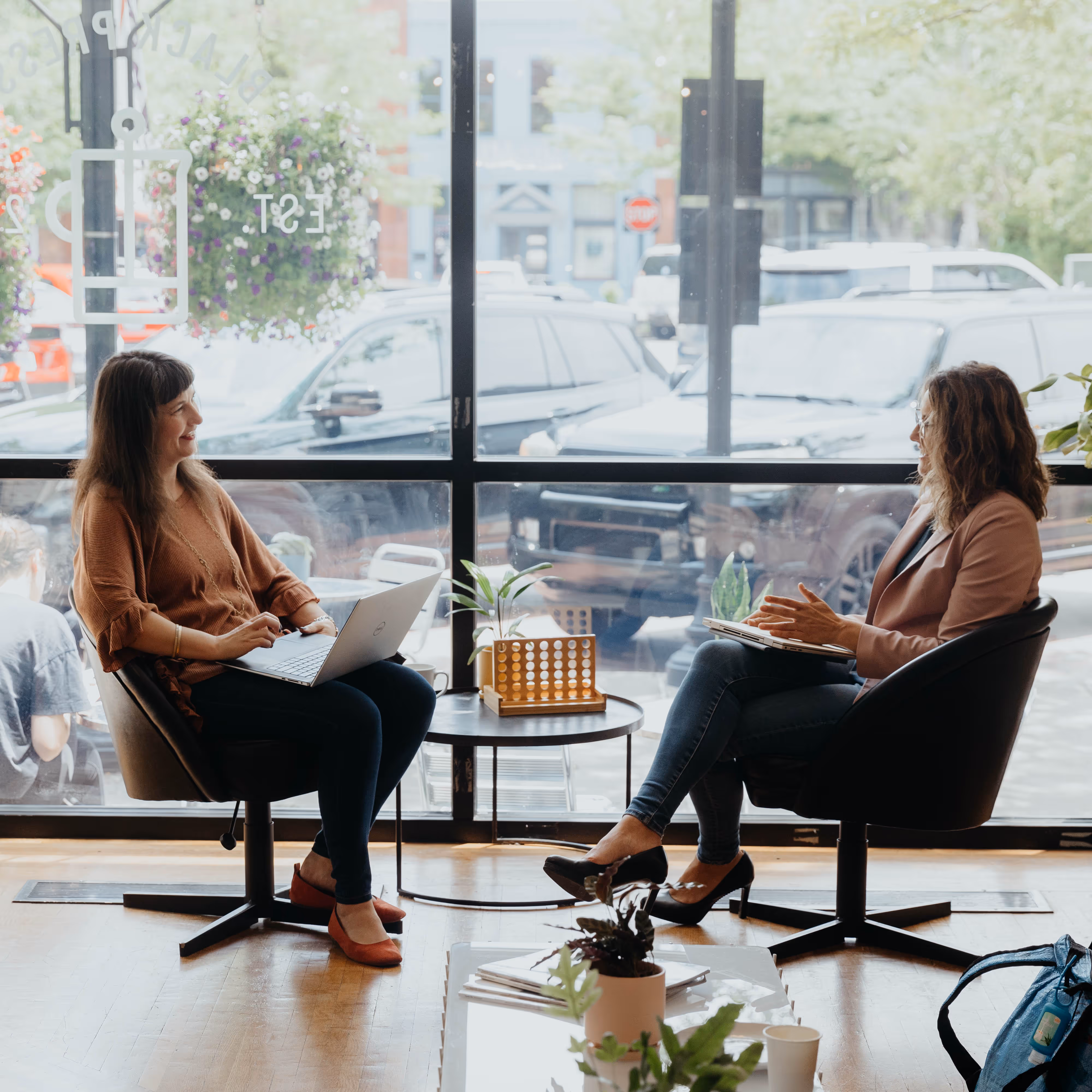Two women sitting in chairs by a large window, engaged in conversation with a laptop and notebook.