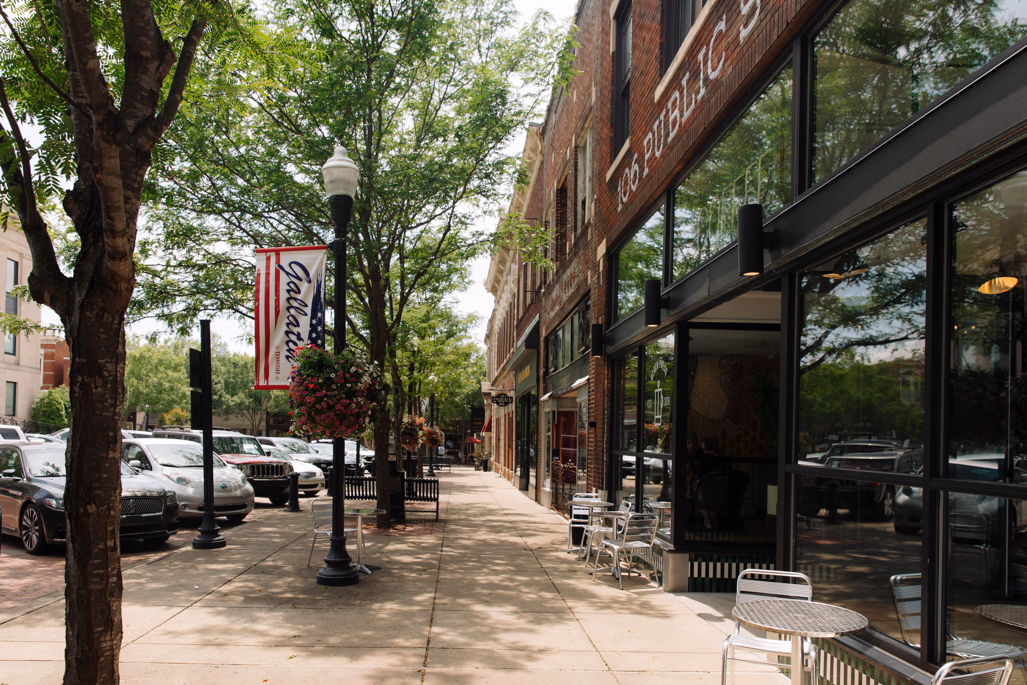 Sunny street in Gallatin, Tennessee, with trees, parked cars, hanging flower baskets, and outdoor café tables.