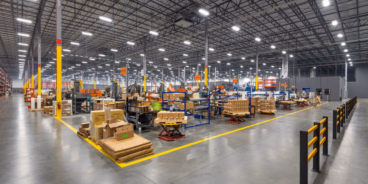 Large, brightly lit warehouse with multiple pallets of cardboard boxes and industrial shelving units organized in rows.