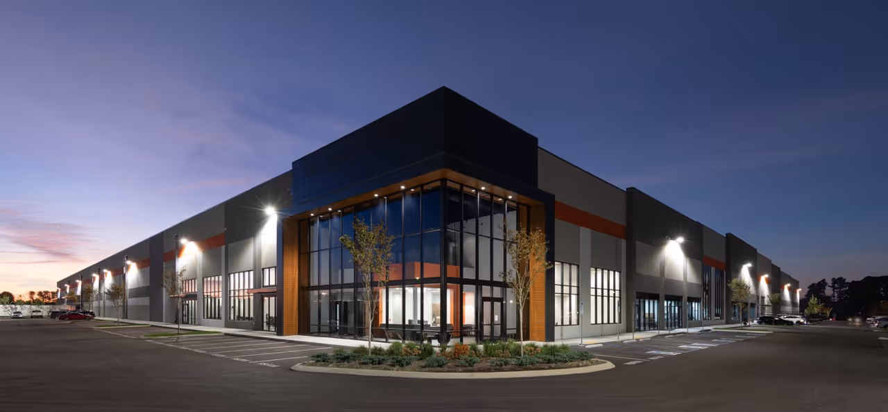 Modern industrial warehouse building with large glass entrance and surrounding parking lot at dusk.
