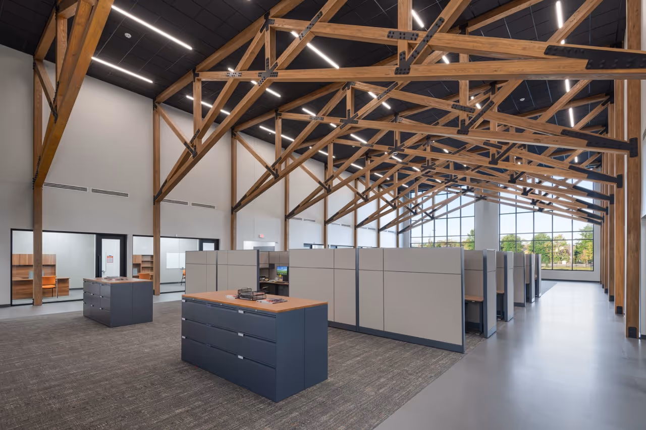 Modern office space with wooden beam ceiling, rows of gray cubicles, filing cabinets, and large window with greenery outside.