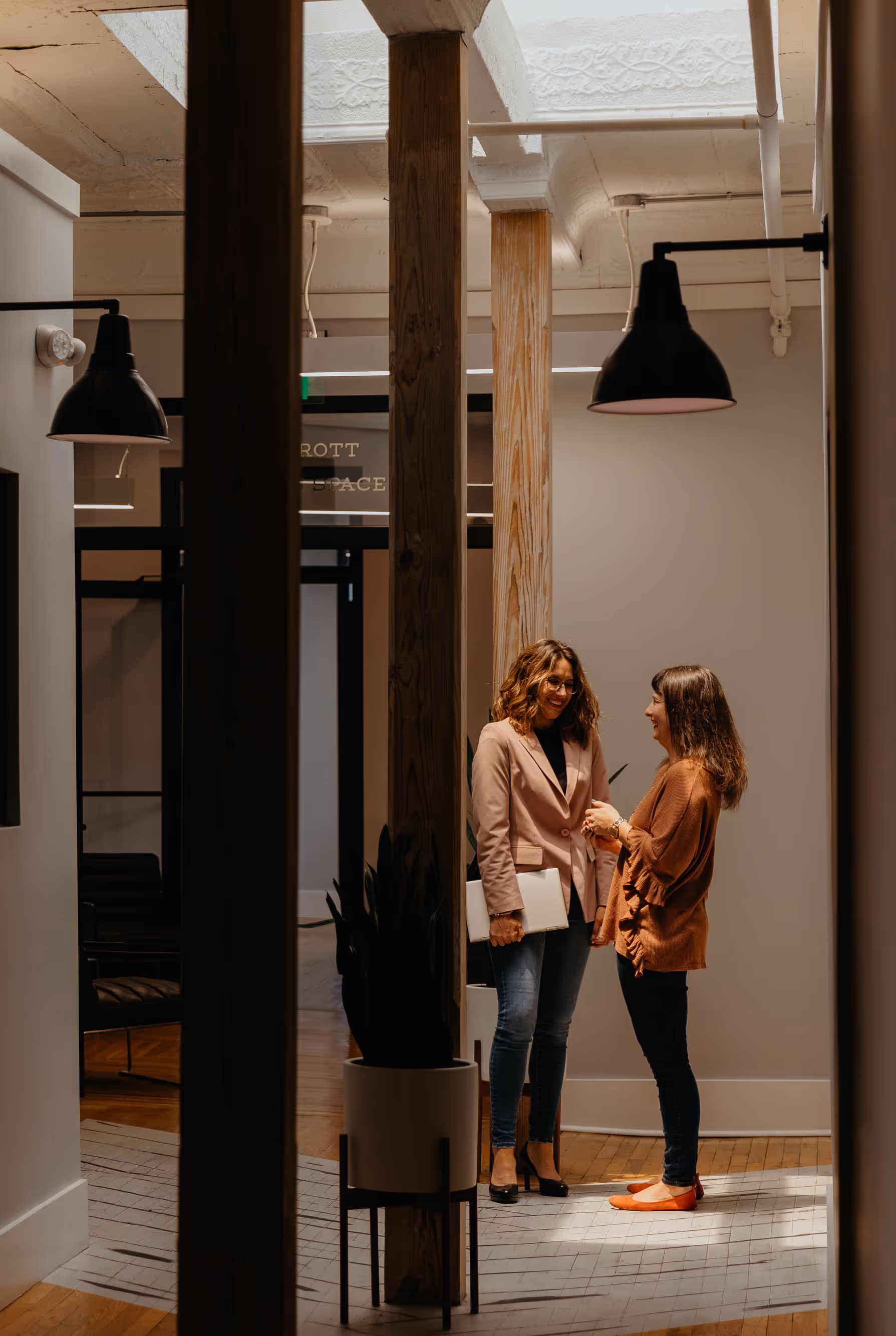 Man in a black blazer reviewing architectural blueprints on a wooden table in a modern room with yellow chairs and green plants.