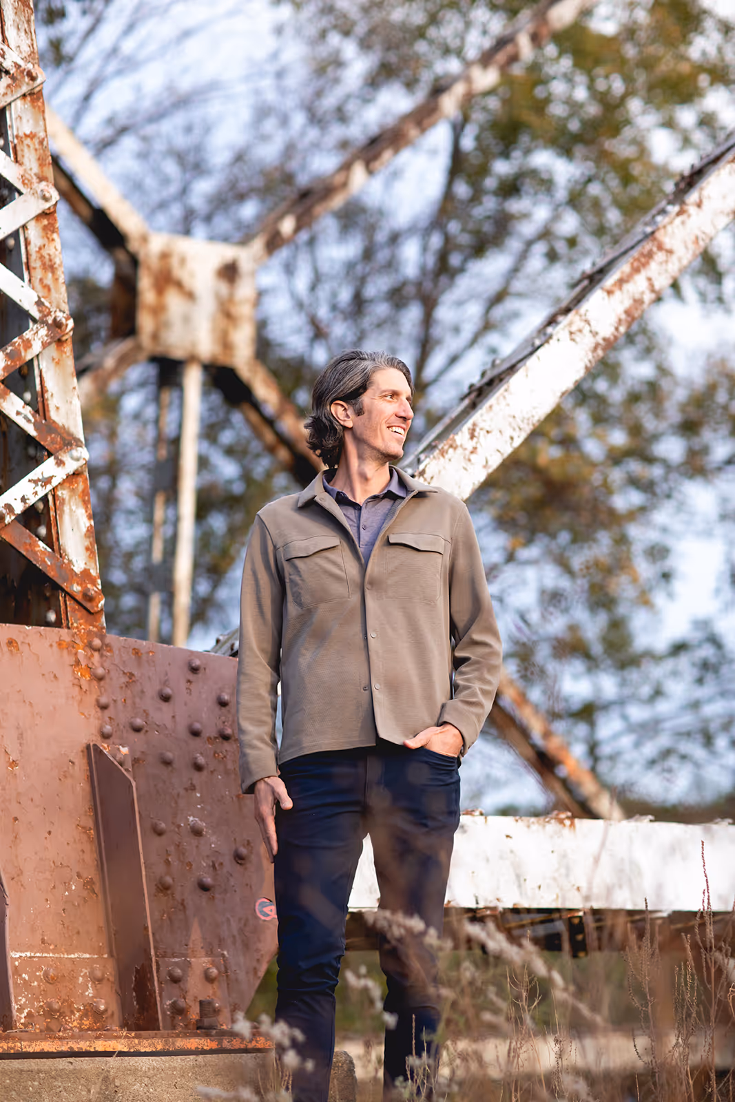 Man in a beige jacket standing outdoors near a rusted metal structure with trees in the background.