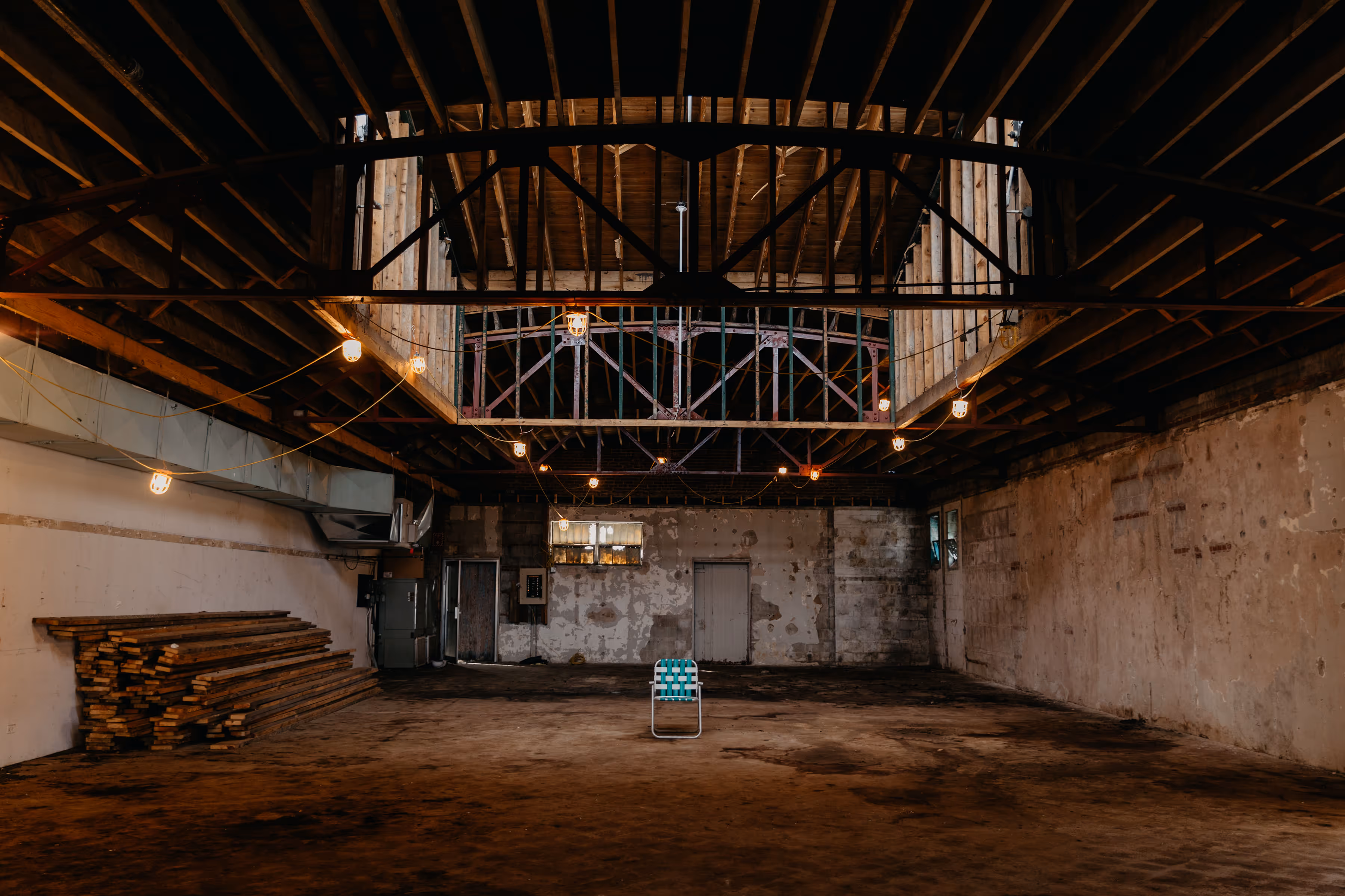 Empty industrial warehouse with exposed wooden beams, hanging string lights, and a single green-and-white striped folding chair in the center.