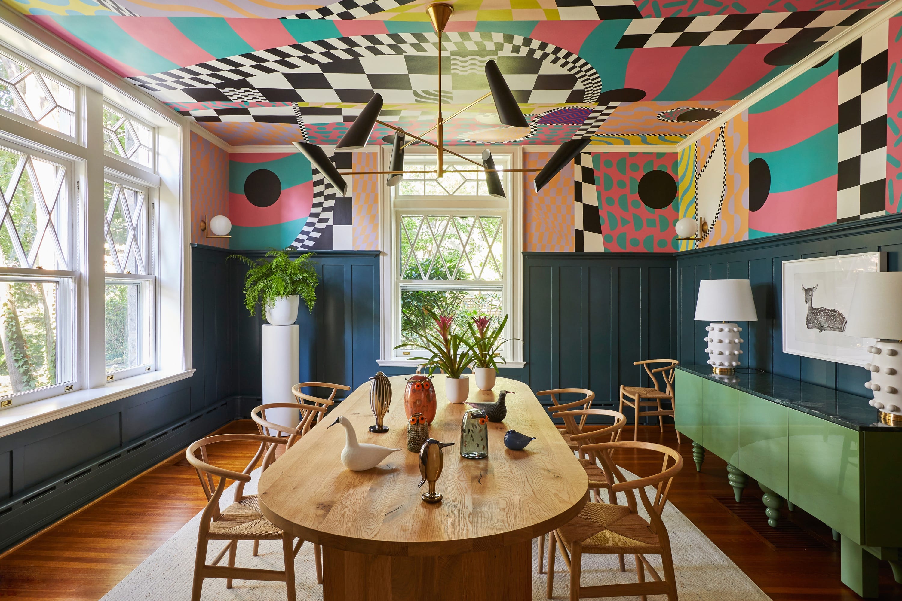 Dining room with a long wooden table surrounded by light wooden chairs, colorful geometric patterns on the upper walls and ceiling, dark blue wainscoting, large windows, and modern light fixtures hanging from the ceiling.