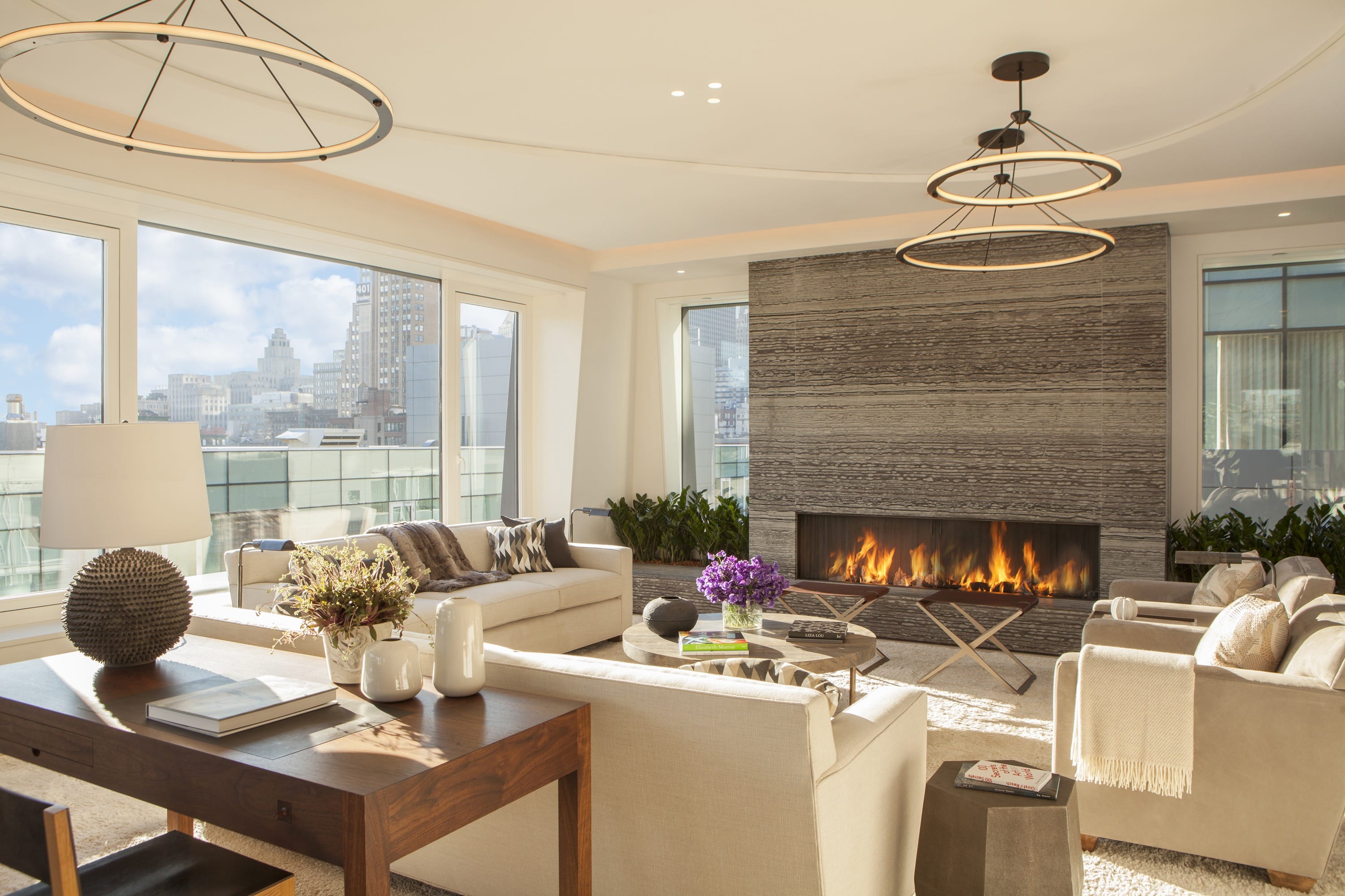 Bright modern living room with large windows showing cityscape, beige sofas, a fireplace with flames, and contemporary round chandeliers.
