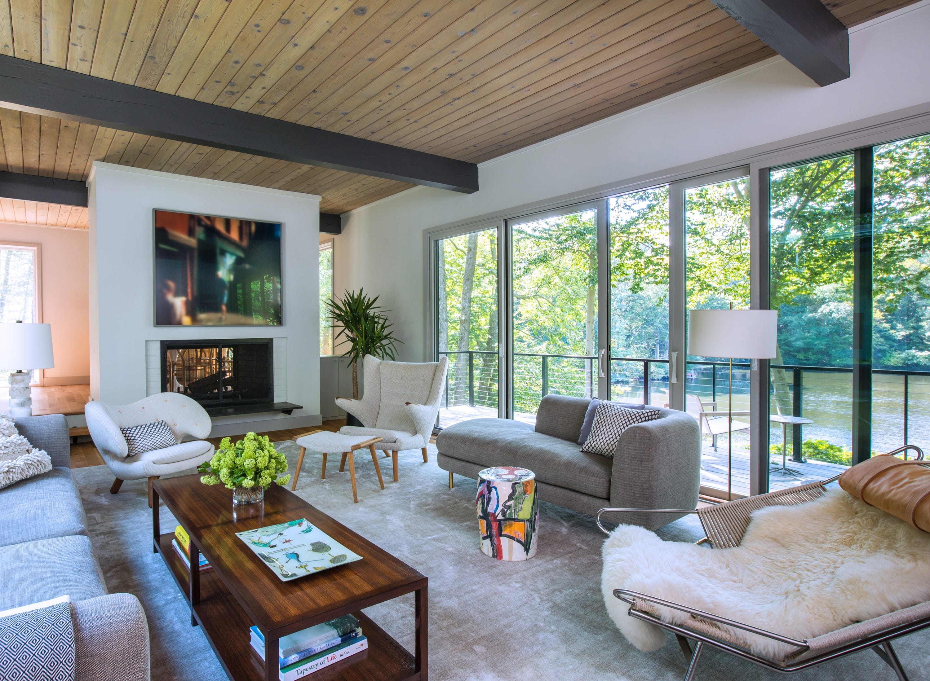 Bright modern living room with large glass sliding doors overlooking a lake, featuring gray and white furniture, a wooden coffee table, and a ceiling with exposed beams.
