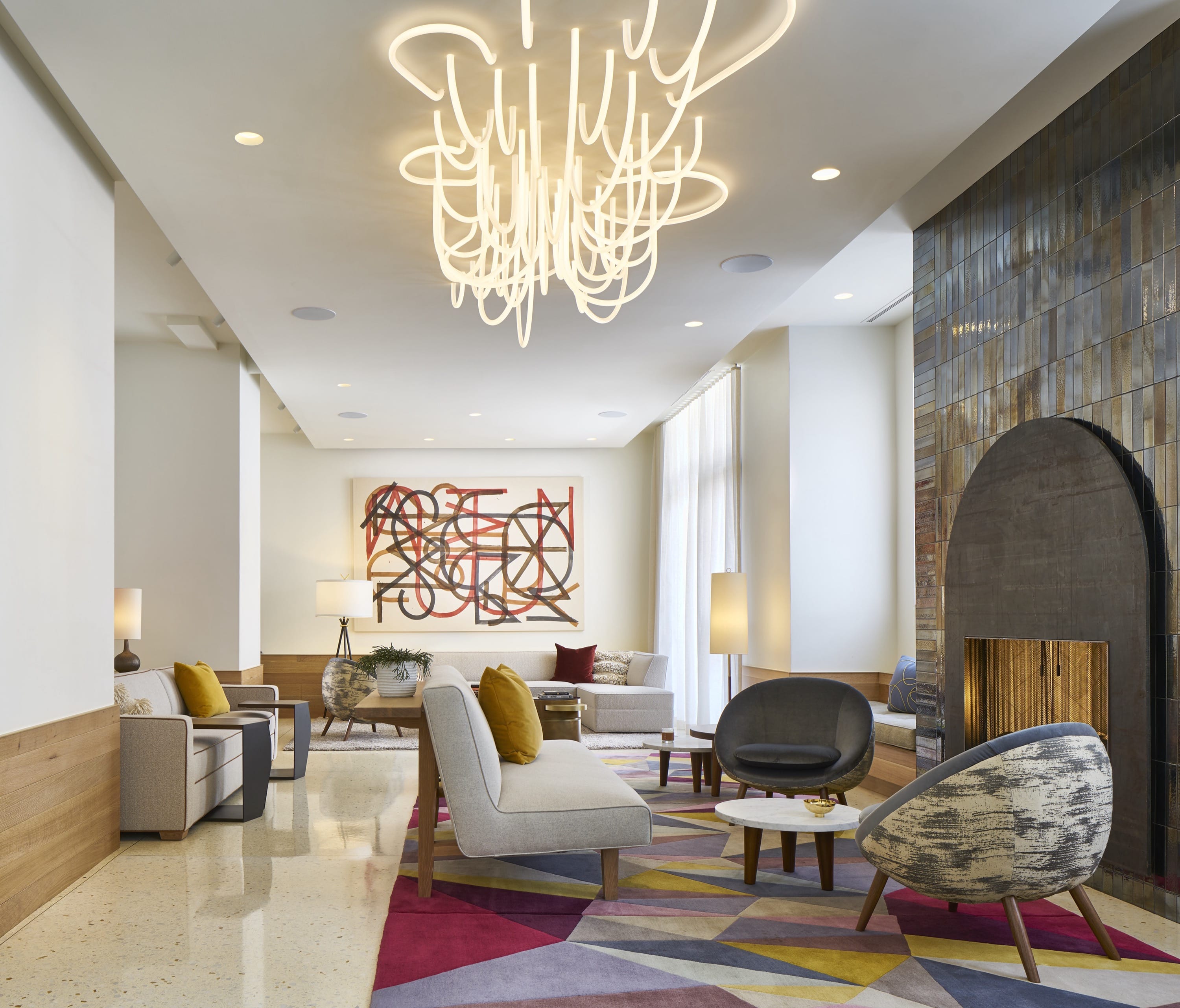 Modern lounge with abstract chandelier, geometric rug, neutral sofas, colorful cushions, and a tiled fireplace.