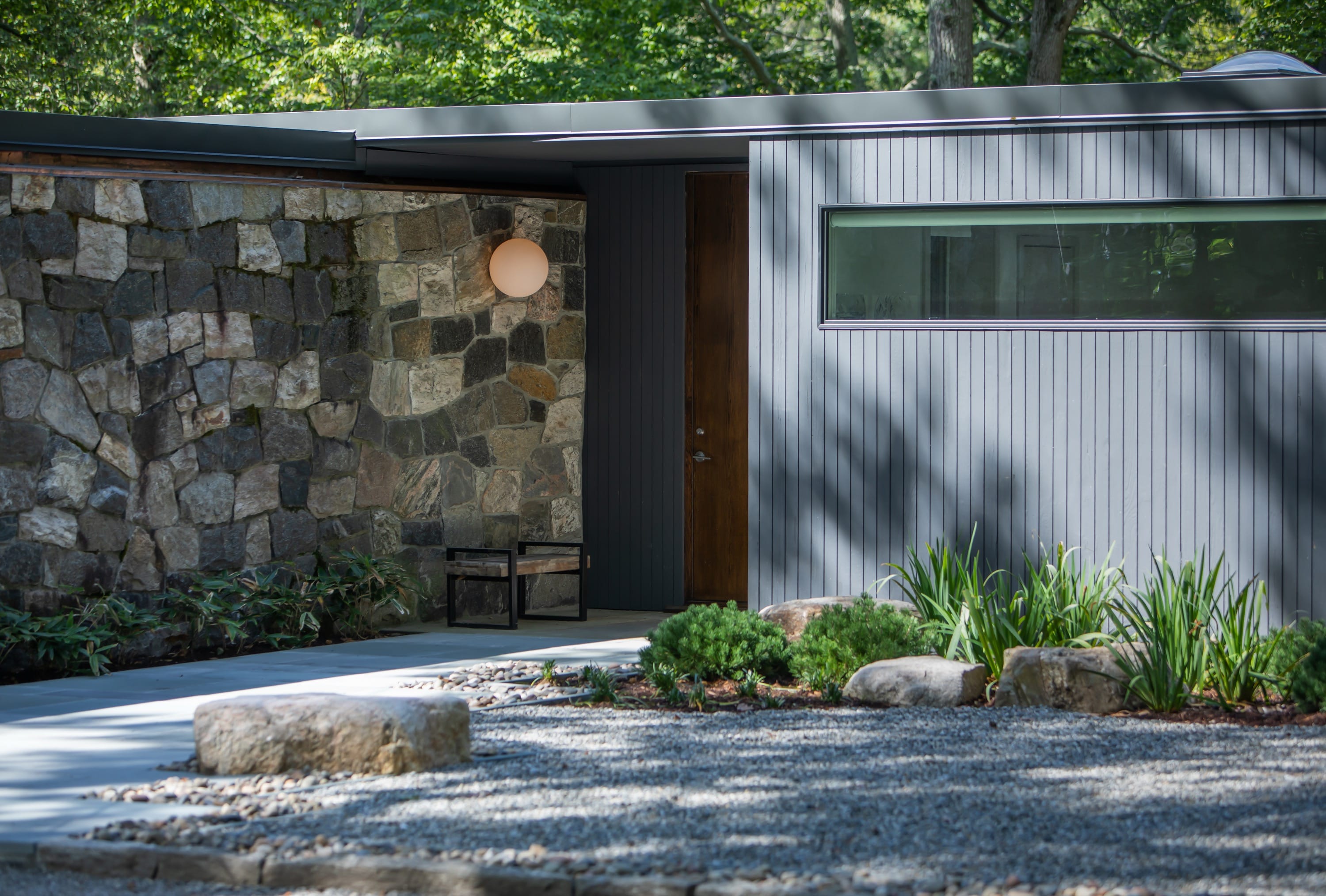 Modern home exterior with stone wall, gray vertical siding, a wooden door, gravel yard, and green shrubs.