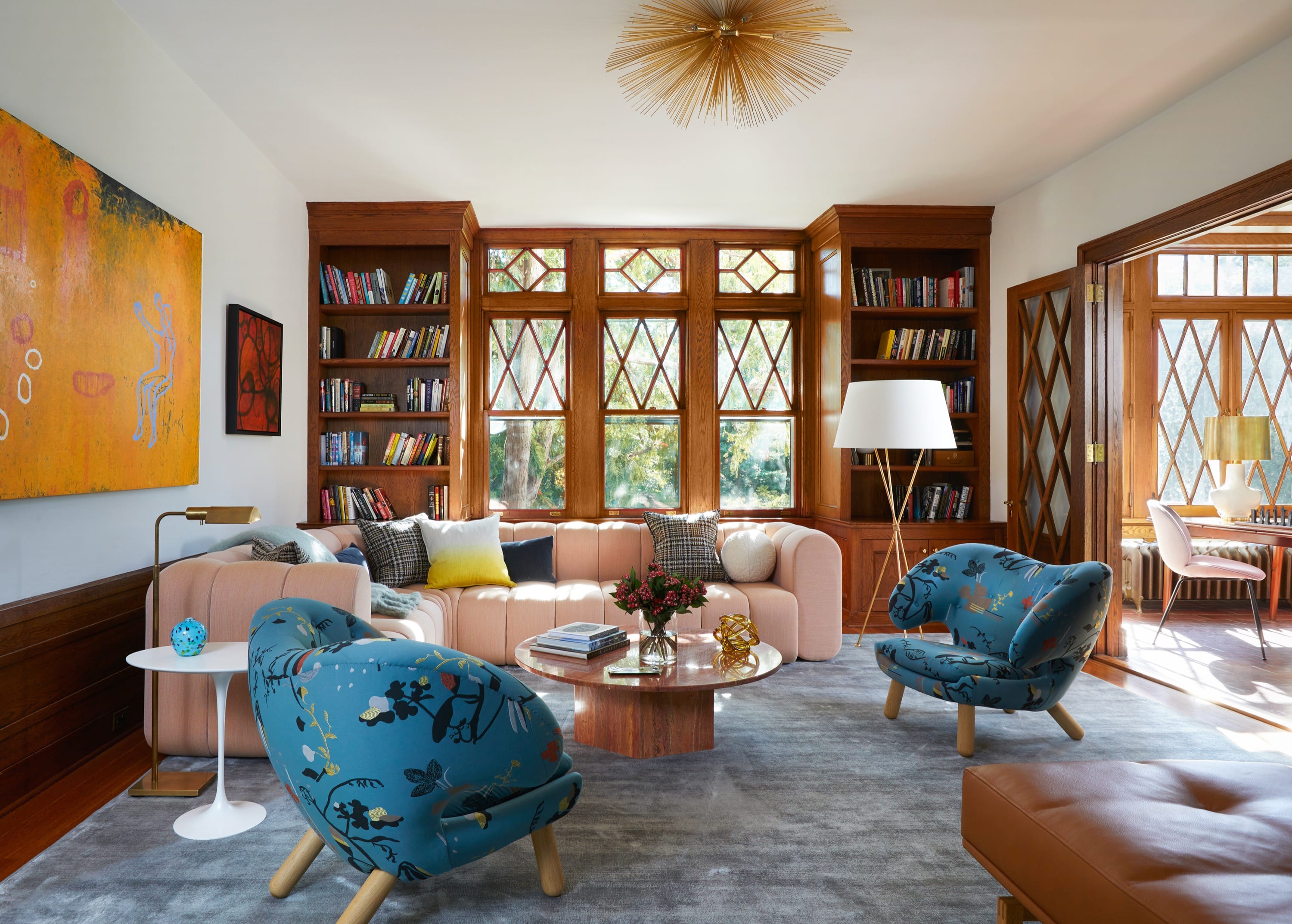 Bright living room with a curved pink sofa, two blue floral armchairs, wooden bookshelves, large windows, and colorful abstract paintings on the wall.