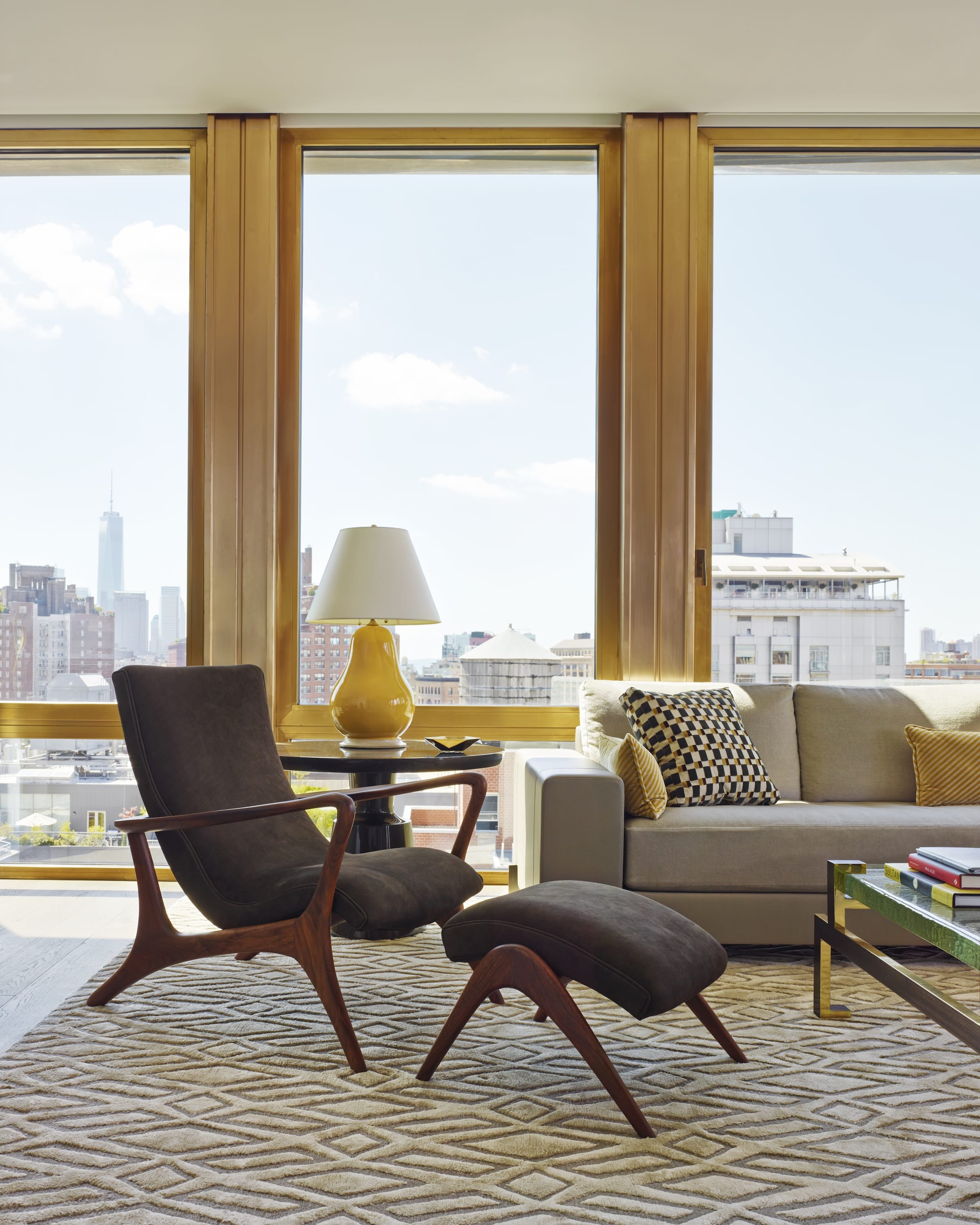 Modern living room with a wooden lounge chair and ottoman on a patterned rug, beige sofa with decorative pillows, and large windows showing city skyline.