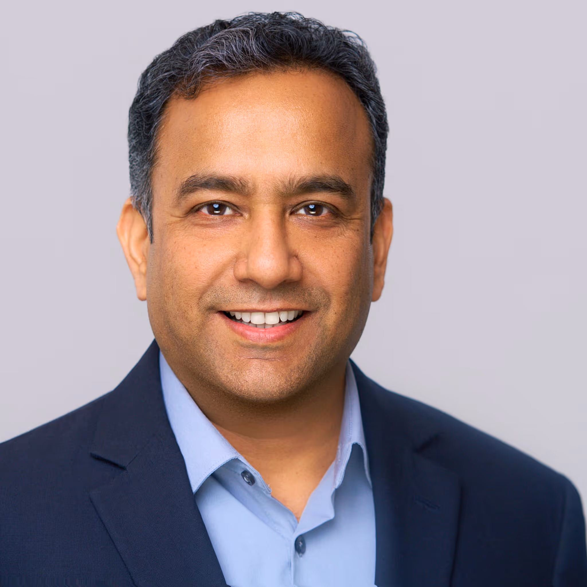 Smiling man in a dark blazer and light blue shirt against a plain light gray background.