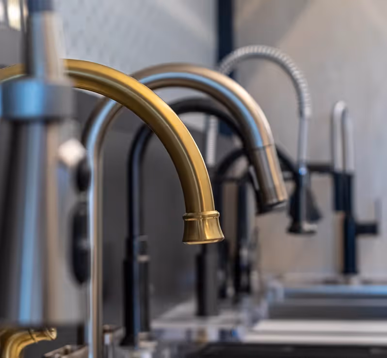Row of kitchen faucets in matte black, brass, and stainless finishes on a white countertop display.