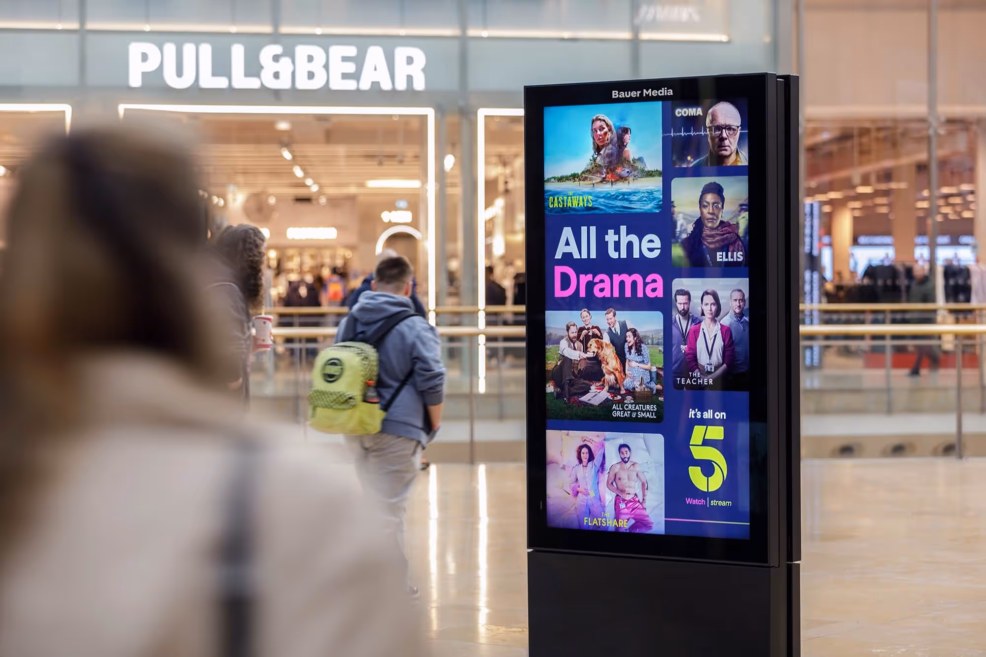 Bauer Media Outdoor digital advertising screen inside a shopping mall displays a Channel 5 ad promoting drama series. 