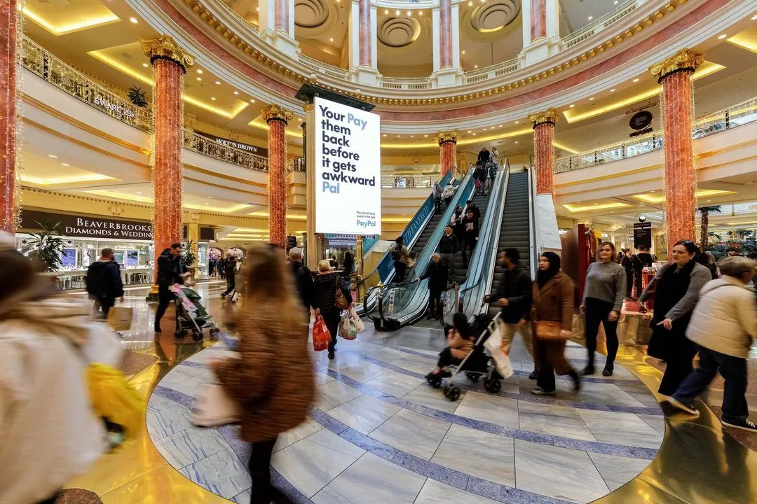 Bauer Media Outdoor digital advertising screen in a shopping mall atrium displays a bold PayPal message: “Pay them back before it gets awkward, Pal,” as shoppers move around escalators and retail stores.