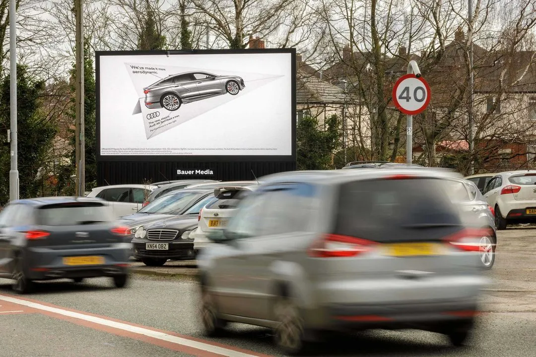 Bauer Media Outdoor roadside digital billboard displays a sleek Audi ad in a residential area, as cars drive past and queue in traffic beneath a 40 mph speed limit sign.