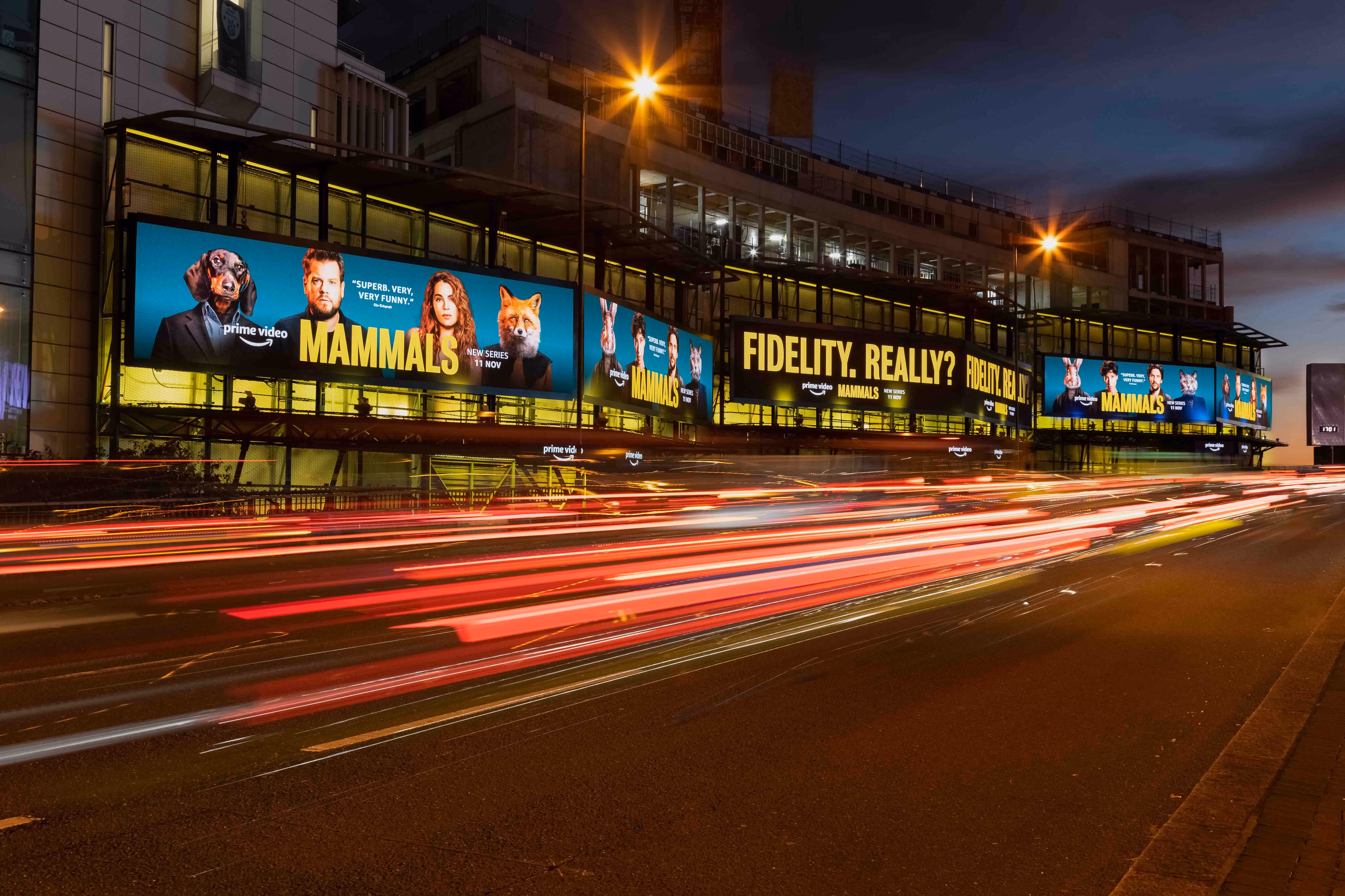Bauer Media Outdoor digital billboards on a city building display an Amazon Prime Video campaign for the series “Mammals,” with bold text reading “FIDELITY, REALLY? RUTH?” as traffic streaks by on a busy evening road.